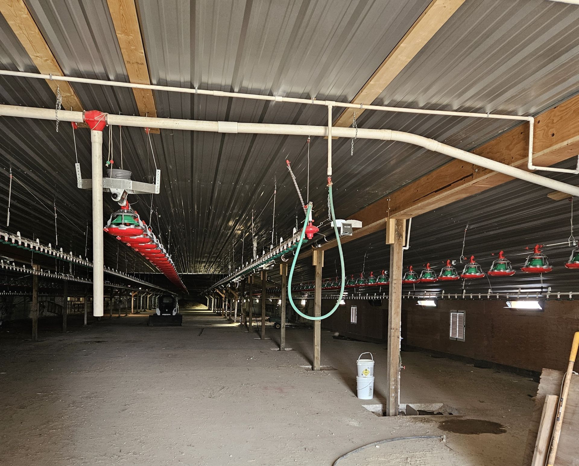 Indoor livestock barn with metal rafters, hanging pipes, hoses, and empty floor.