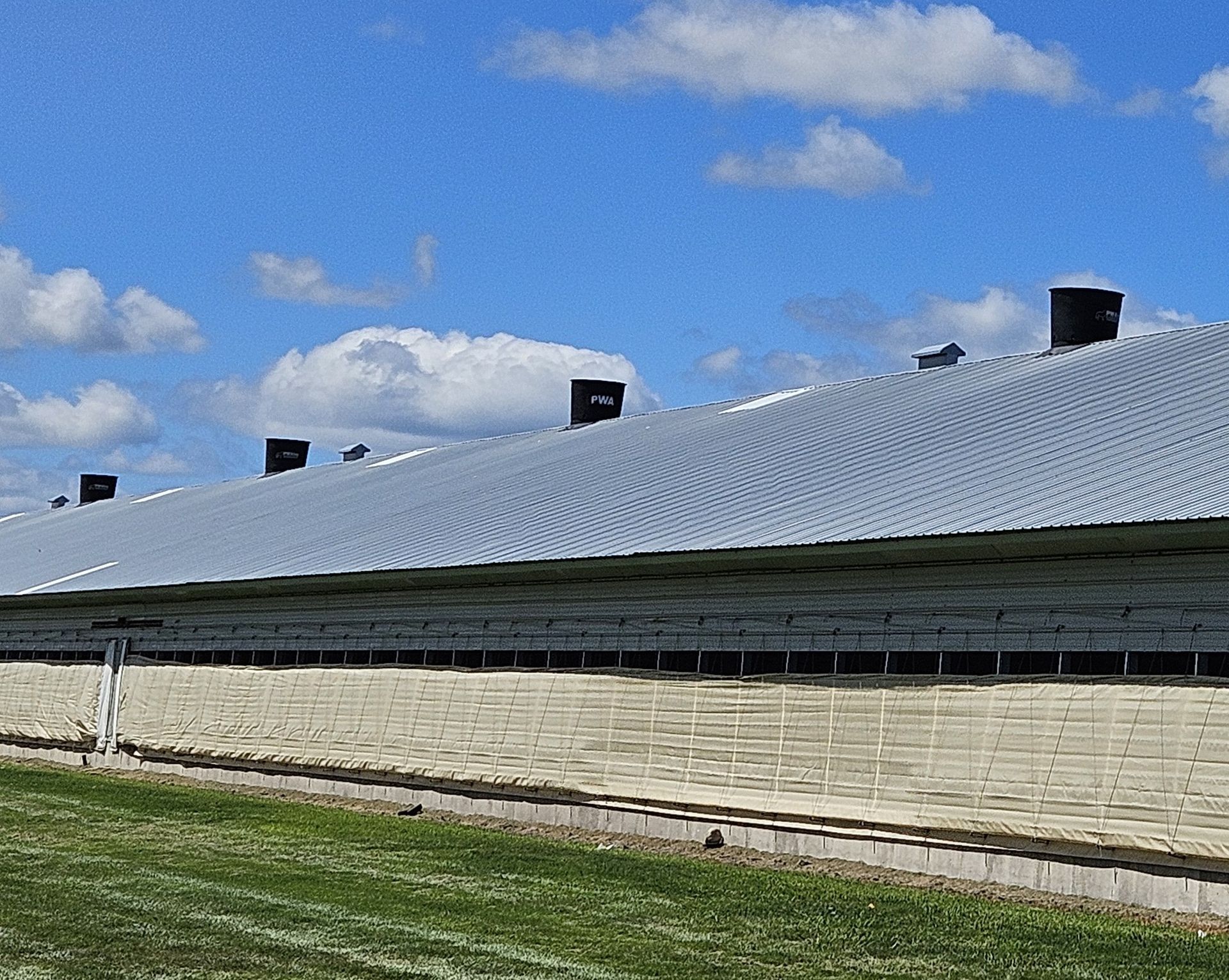 Large silver-roofed barn or industrial building under a blue sky with clouds and grassy foreground