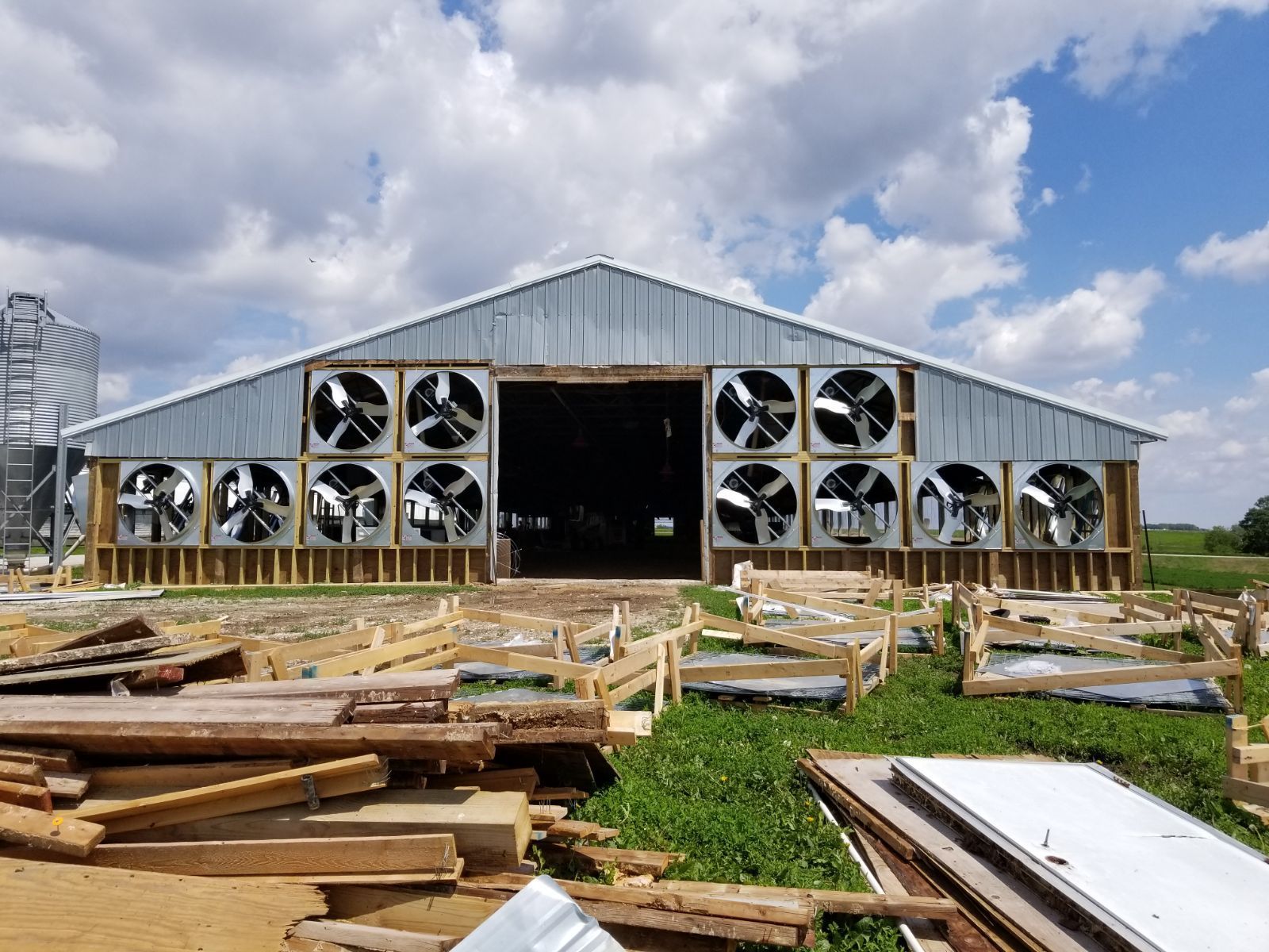 Large barn under construction with metal siding, open front, and stacks of lumber in the foreground
