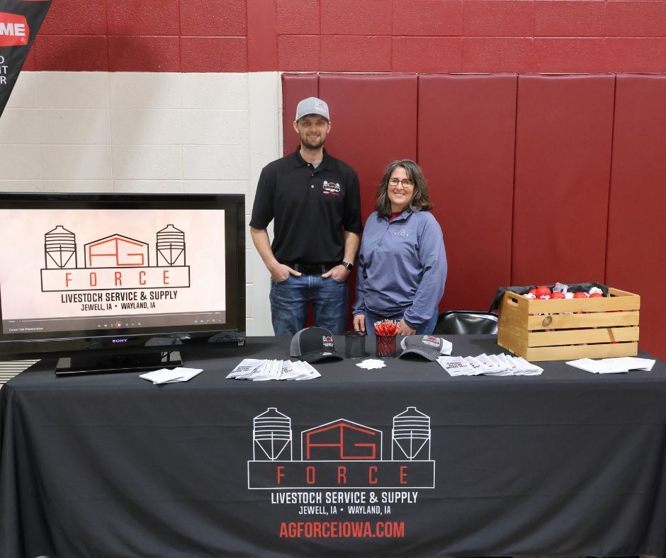 Two people at a vendor booth with a black “Forge” tablecloth and display items on a red wall backdrop