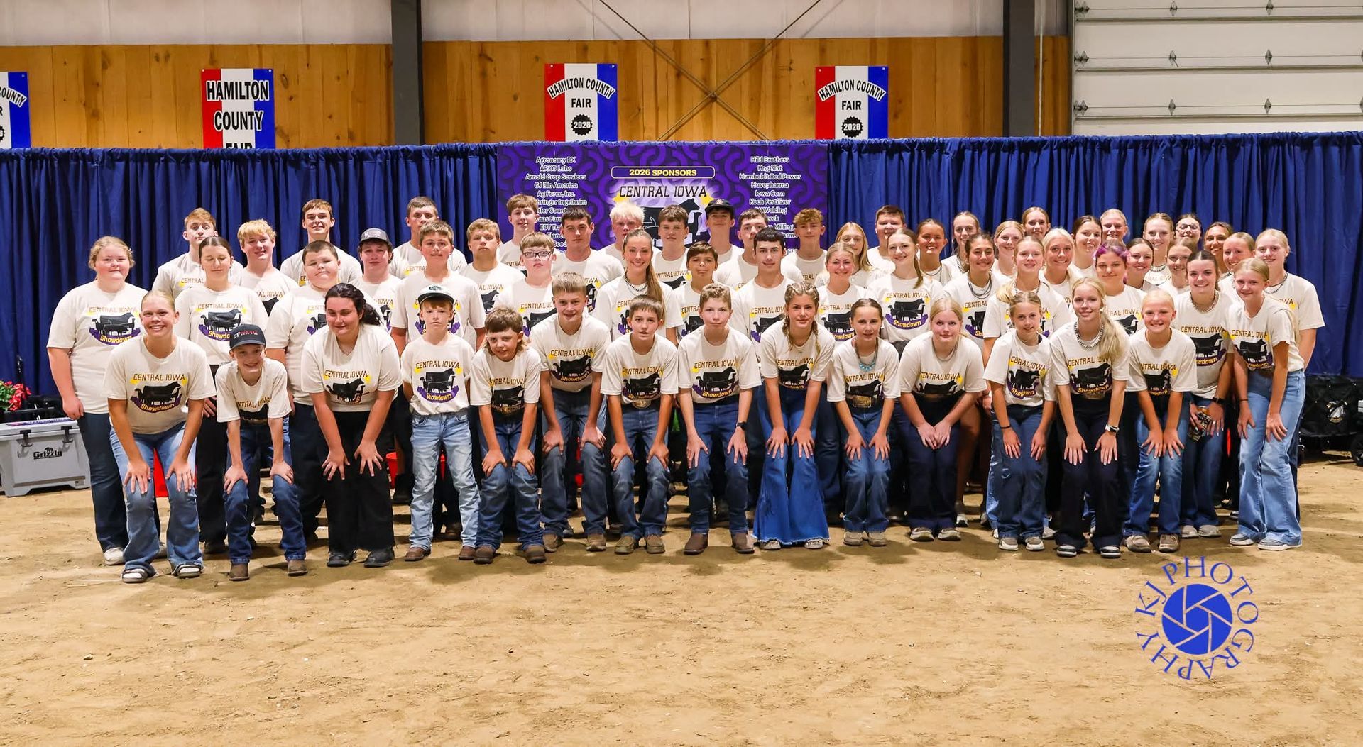 Large group posing for a team photo in a dirt arena with blue and red banners behind them.