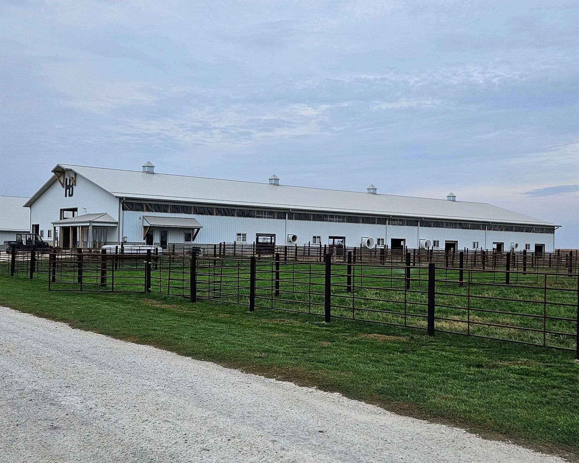 White barn with fenced green pasture under a cloudy sky, beside a gravel road.