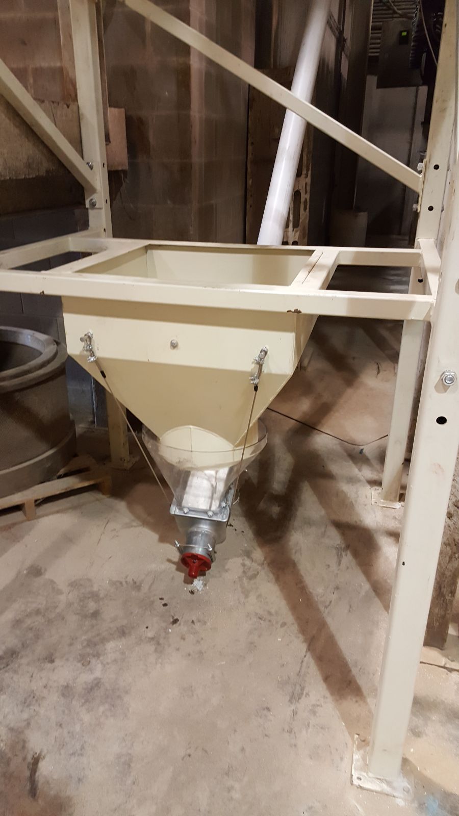White industrial hopper with metal frame and chute in a dusty warehouse or workshop.