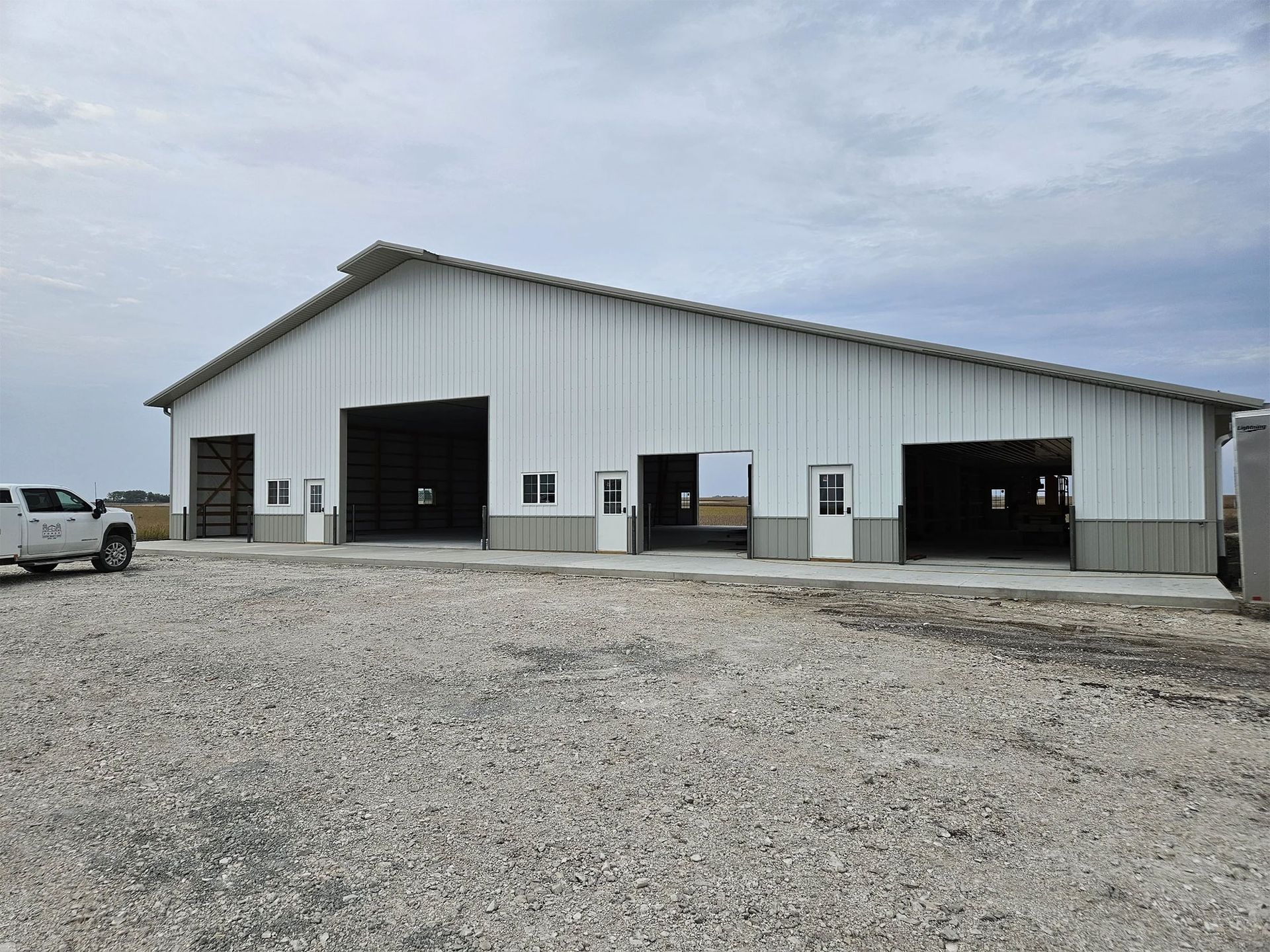 Large white metal warehouse with open garage bays on a gravel lot under a cloudy sky