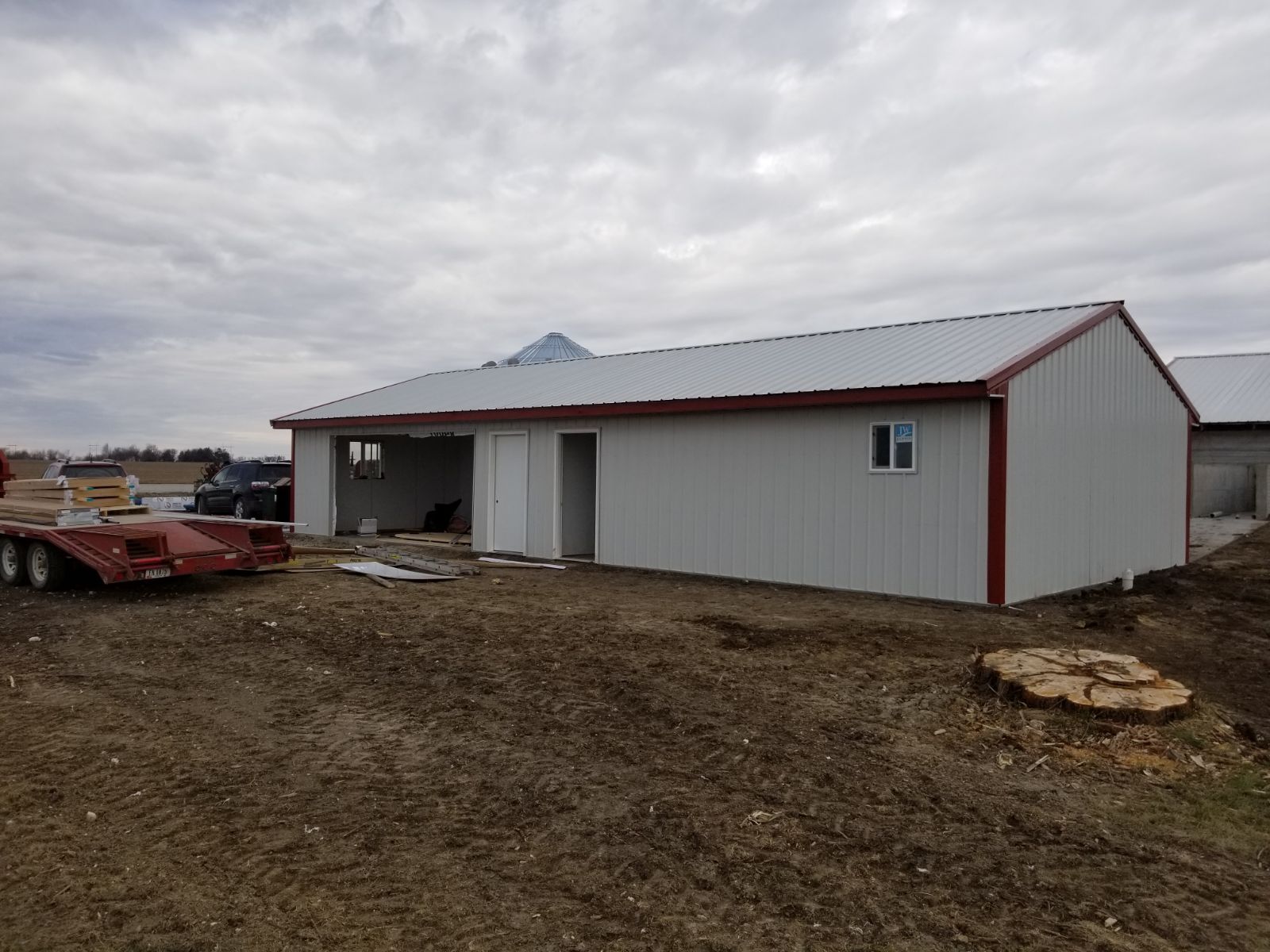 White metal shed with red trim on a dirt lot under cloudy skies, with farm equipment nearby.