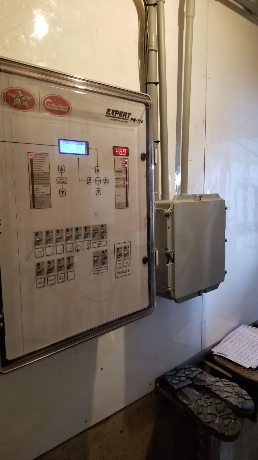 Wall-mounted electrical control panel with lit display and switches next to a gray box in a narrow utility room