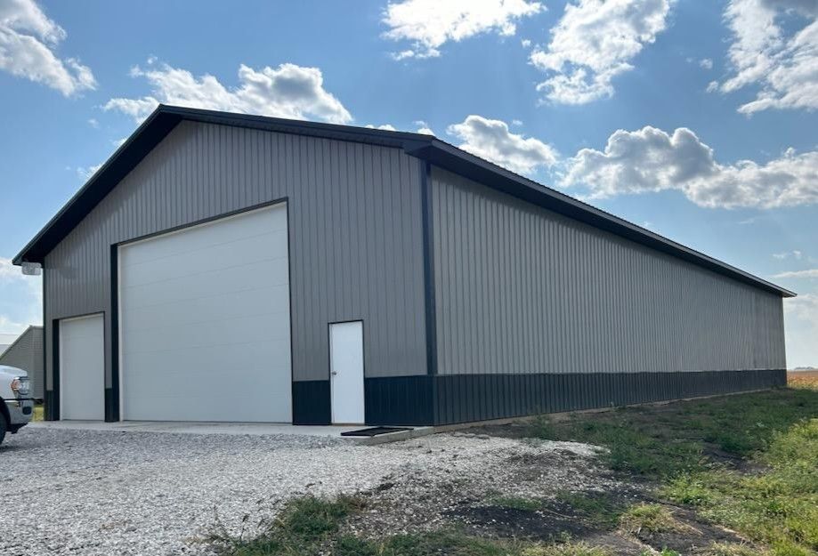 Large gray metal warehouse with a white garage door, set on a gravel lot under a partly cloudy sky