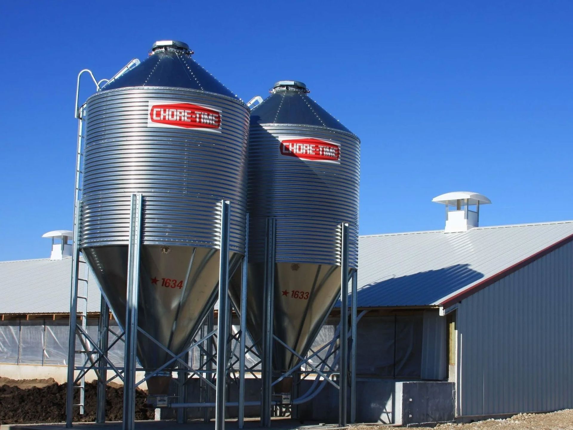 Two large metal grain silos beside a farm building under a clear blue sky