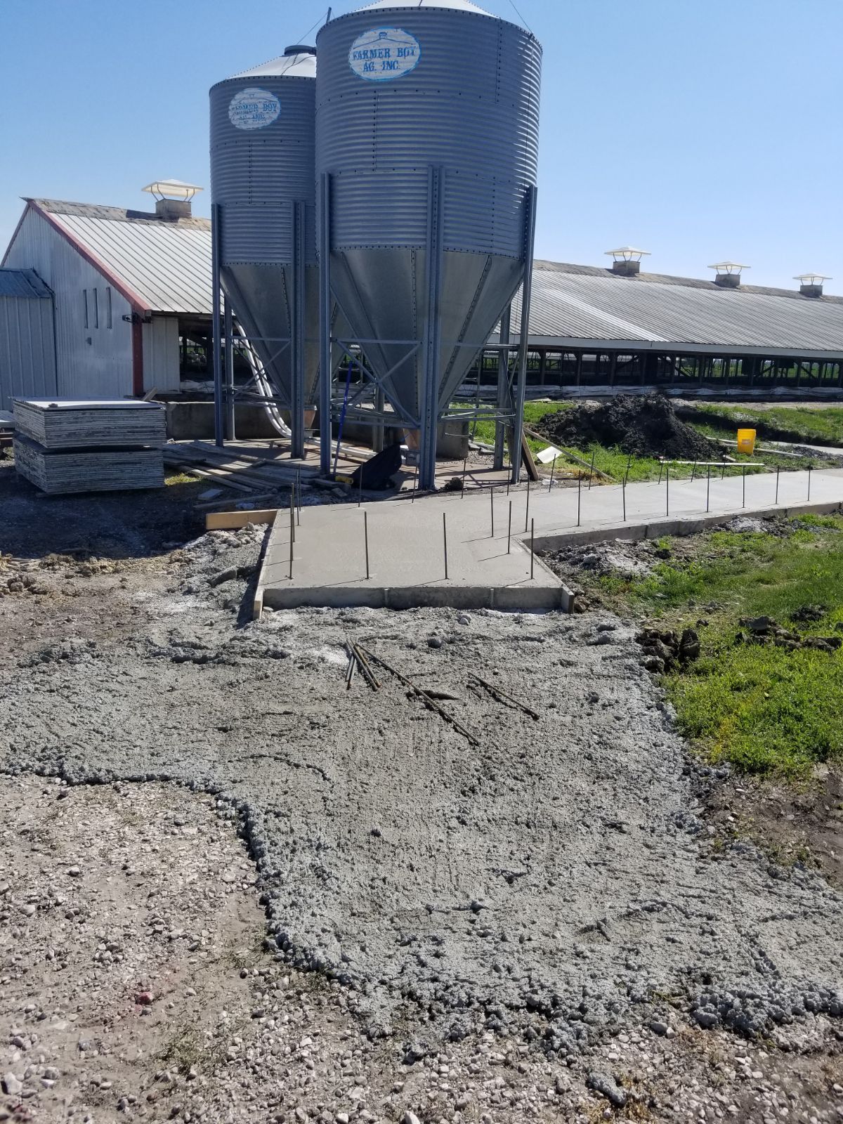 Two metal grain silos beside a farm building, with a gravel path in the foreground under a clear sky.