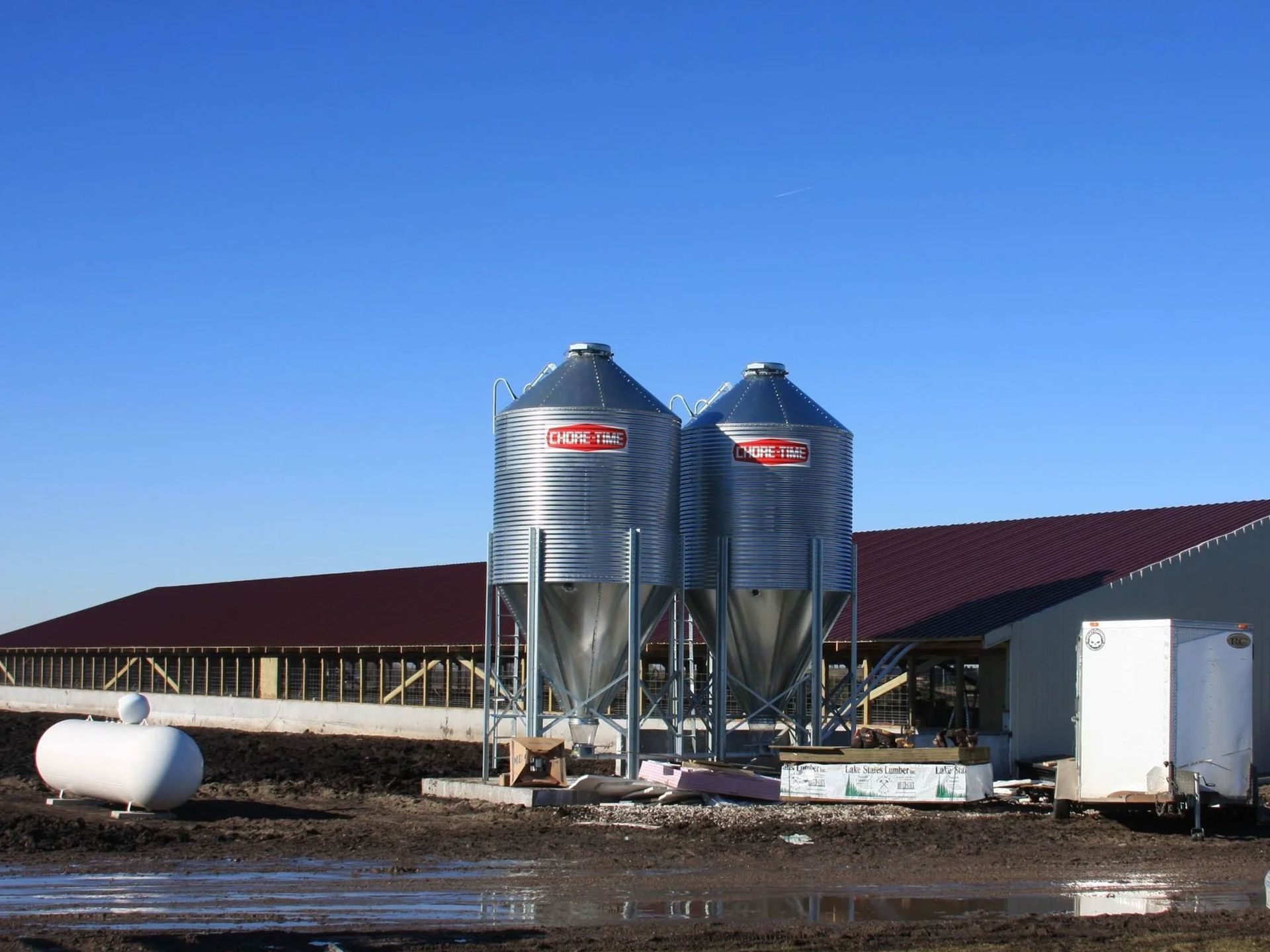Two large metal grain silos beside a red-roofed farm building under a clear blue sky