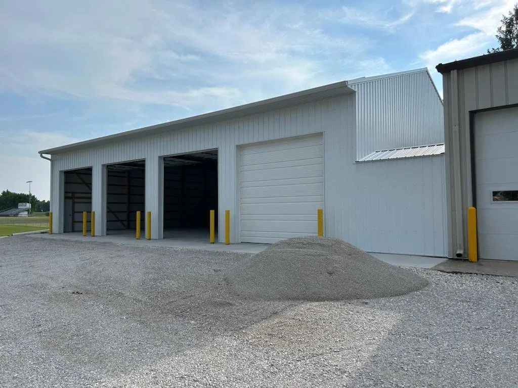 White industrial building with open garage bays, gravel foreground, and yellow safety bollards
