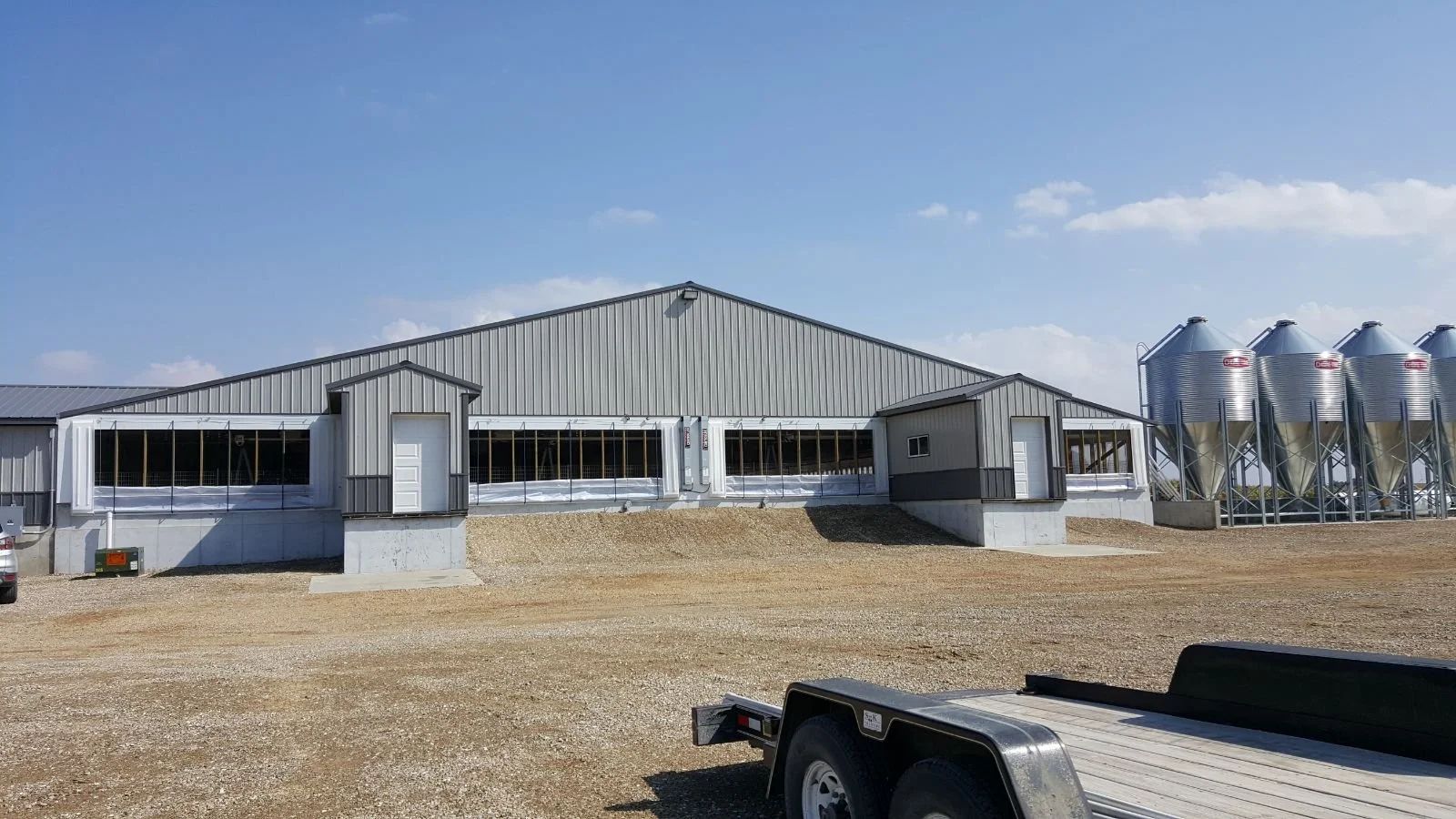 Rural farm building with metal siding and silos on a gravel lot under a clear blue sky
