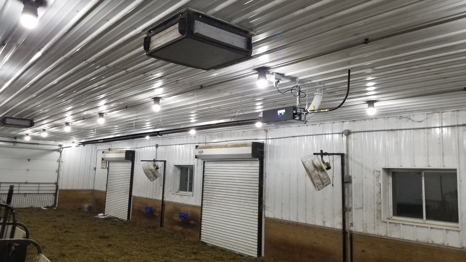 Interior view of a barn showing white corrugated metal walls, ceiling heater, circulation fans, and roll-up doors.
