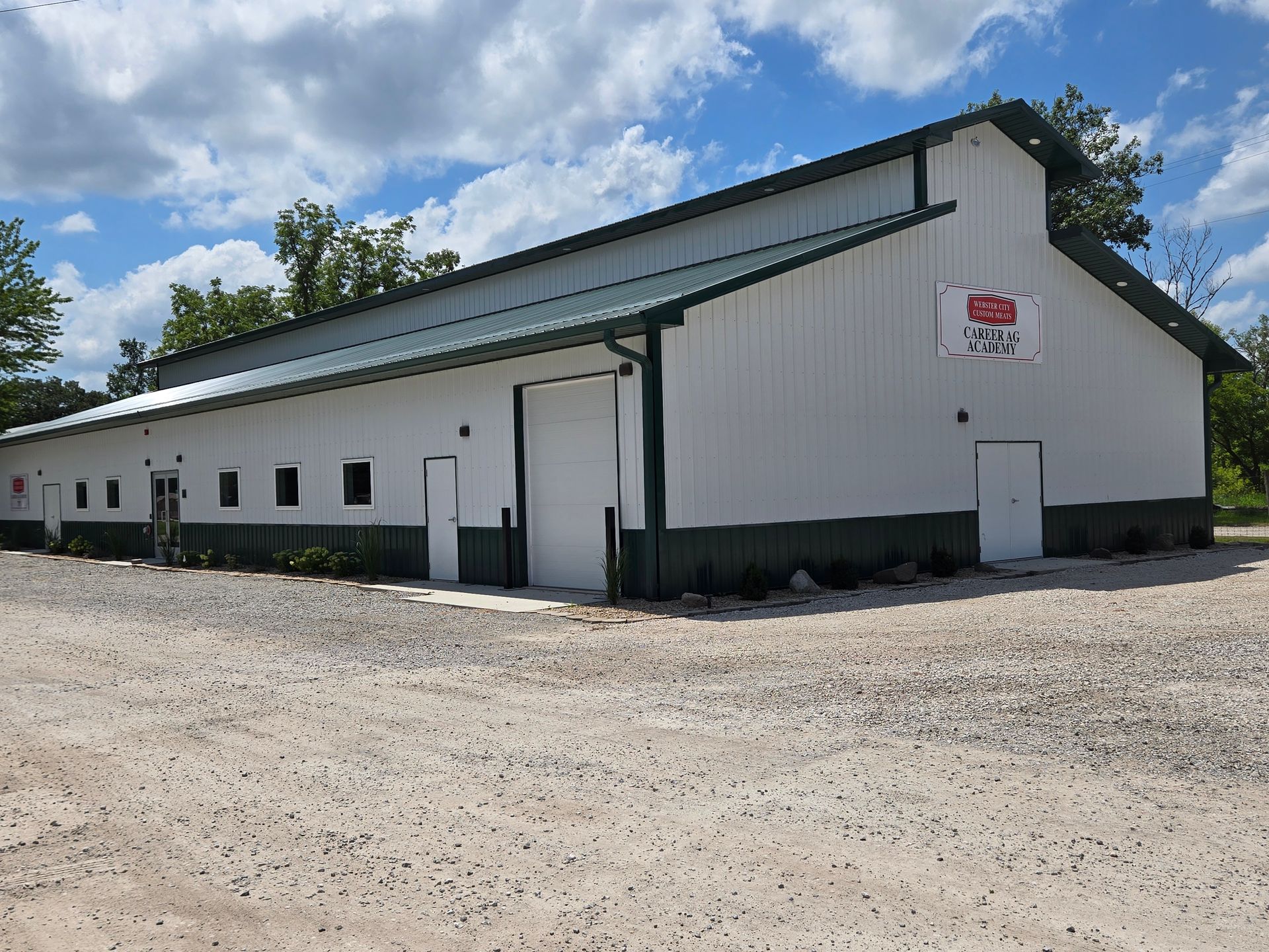 Exterior view of a large white metal building with dark green trim and a sign reading Webster City Custom Meats Career Ag Academy set on a gravel lot.