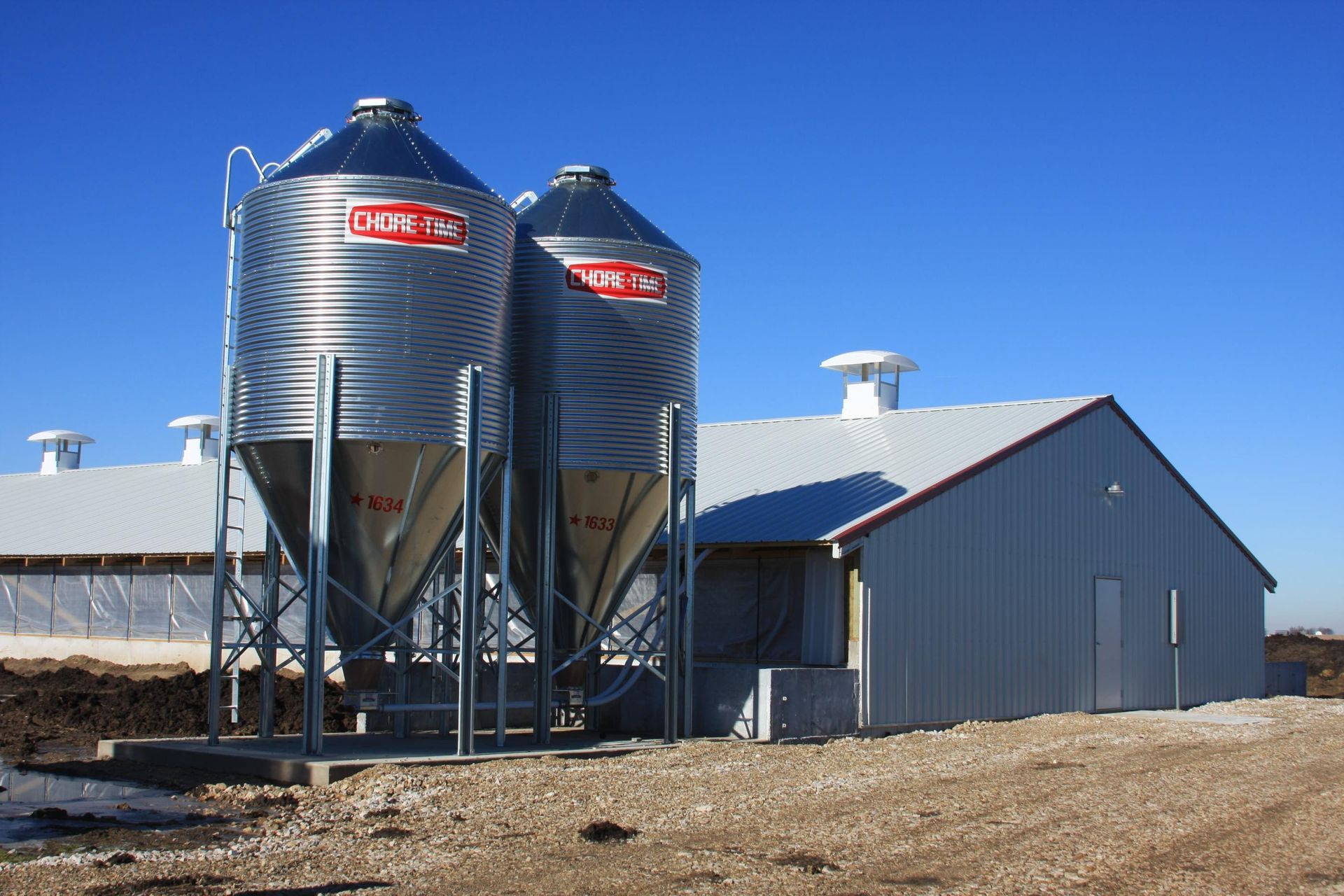 Two Chore-Time feed silos mounted next to a white metal agricultural livestock facility against a clear blue sky.