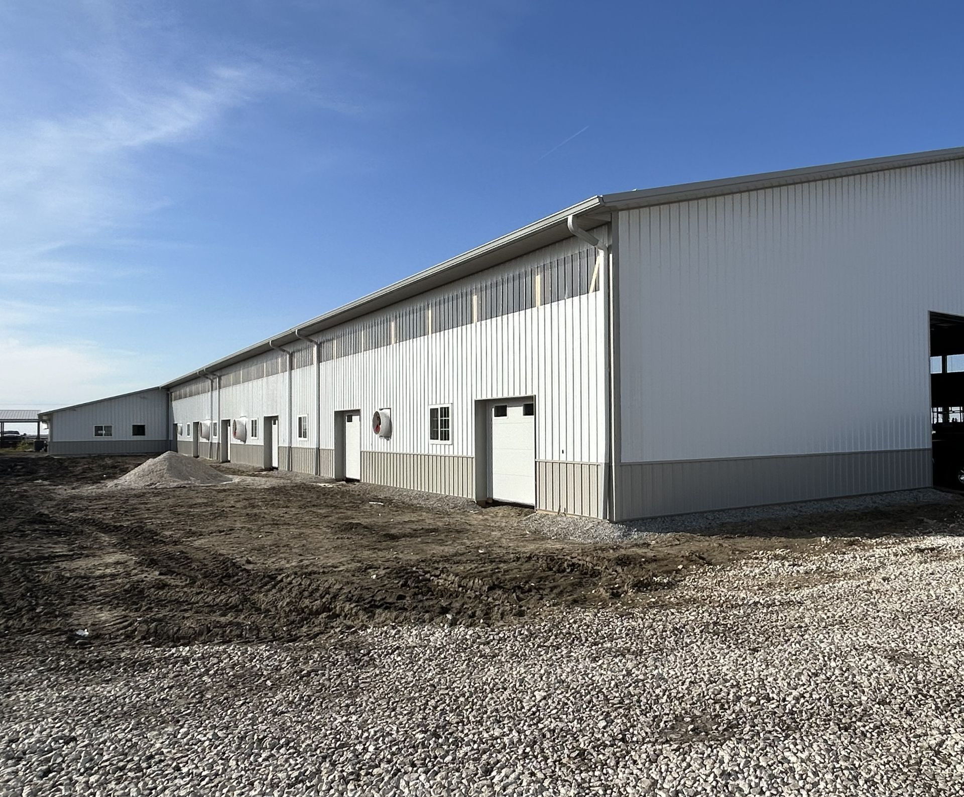 Exterior side view of an extended white metal agricultural building featuring ventilation fans, small entry doors, and a gravel driveway.