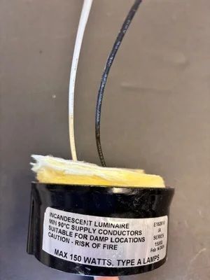 Close-up of a black lamp socket with two exposed wires and a warning label against a beige background