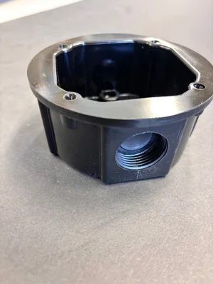 Black plastic electrical box with silver metal rim and threaded side opening on a gray surface