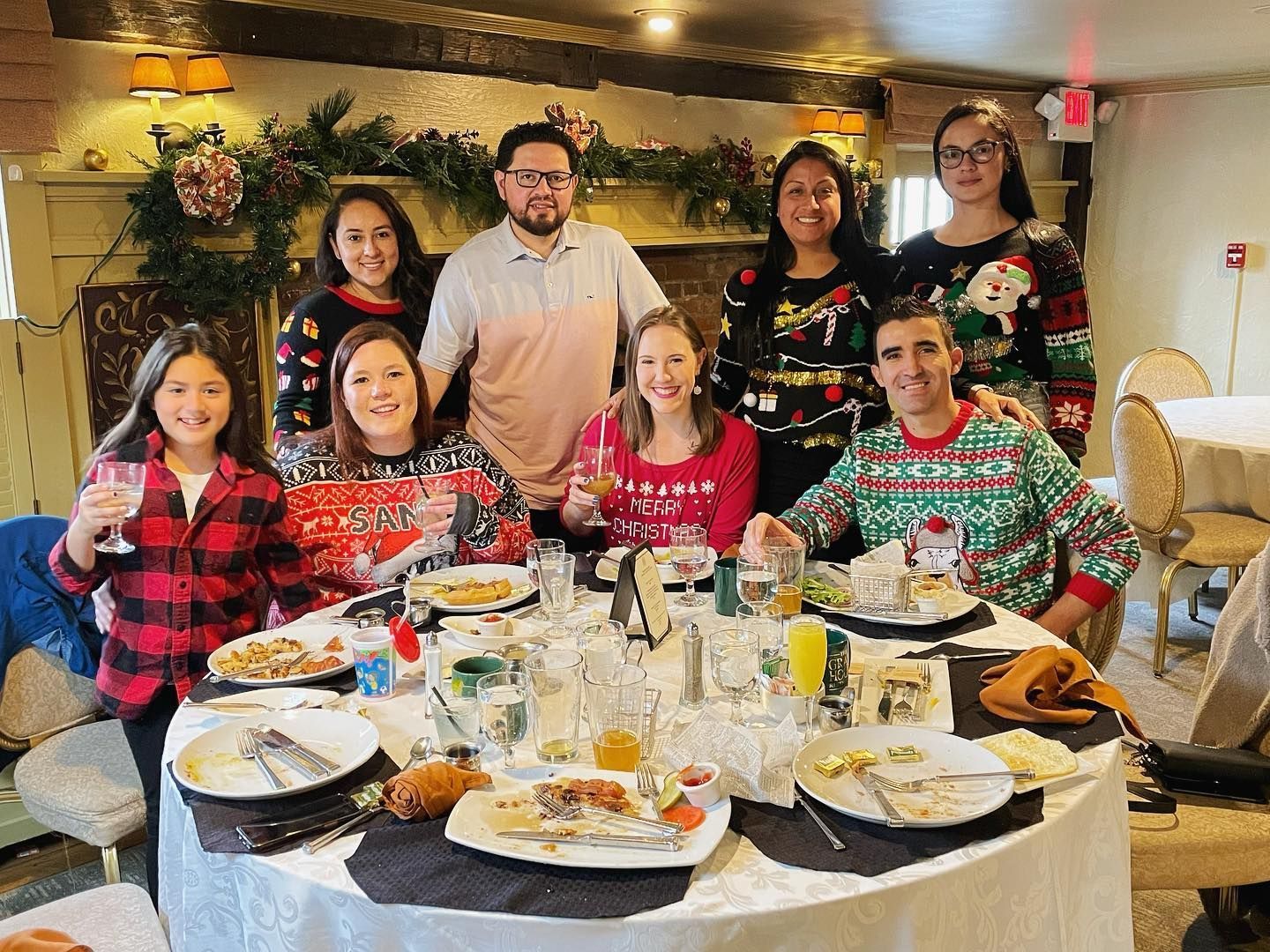 Group of eight seated at a restaurant table with plates, drinks, and holiday sweaters, smiling for a dinner photo