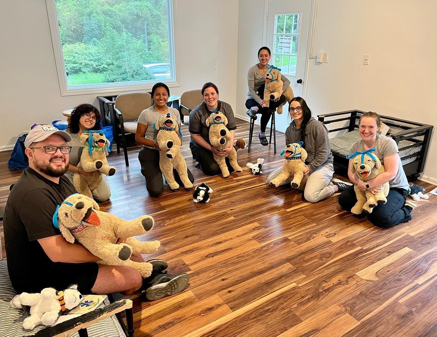 Group seated on hardwood floor in a bright room, each holding a plush toy dog, smiling at the camera.