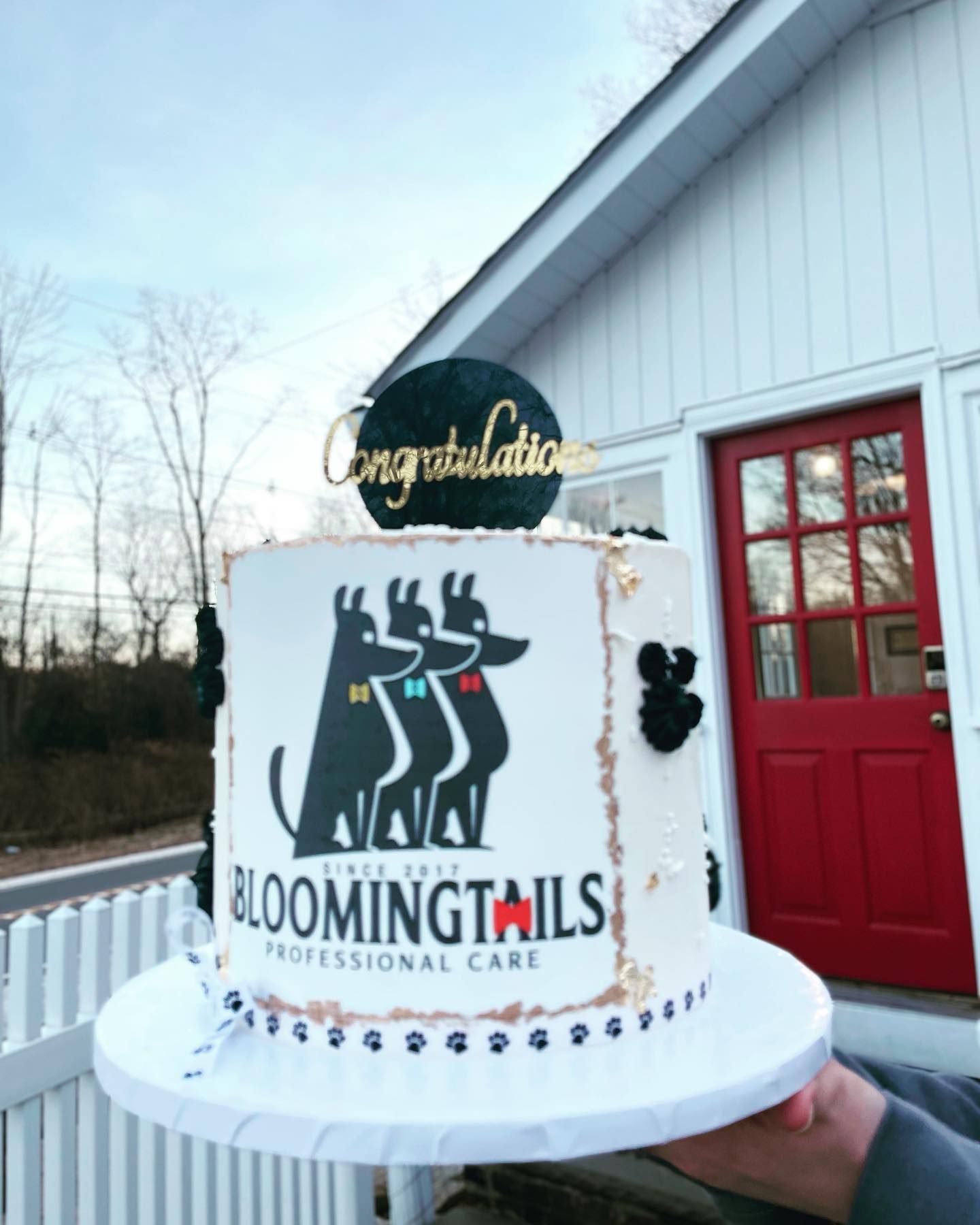 Blooming Tails Professional Care cake with black dog silhouettes and a gold “Congratulations” topper outside a red-doored building