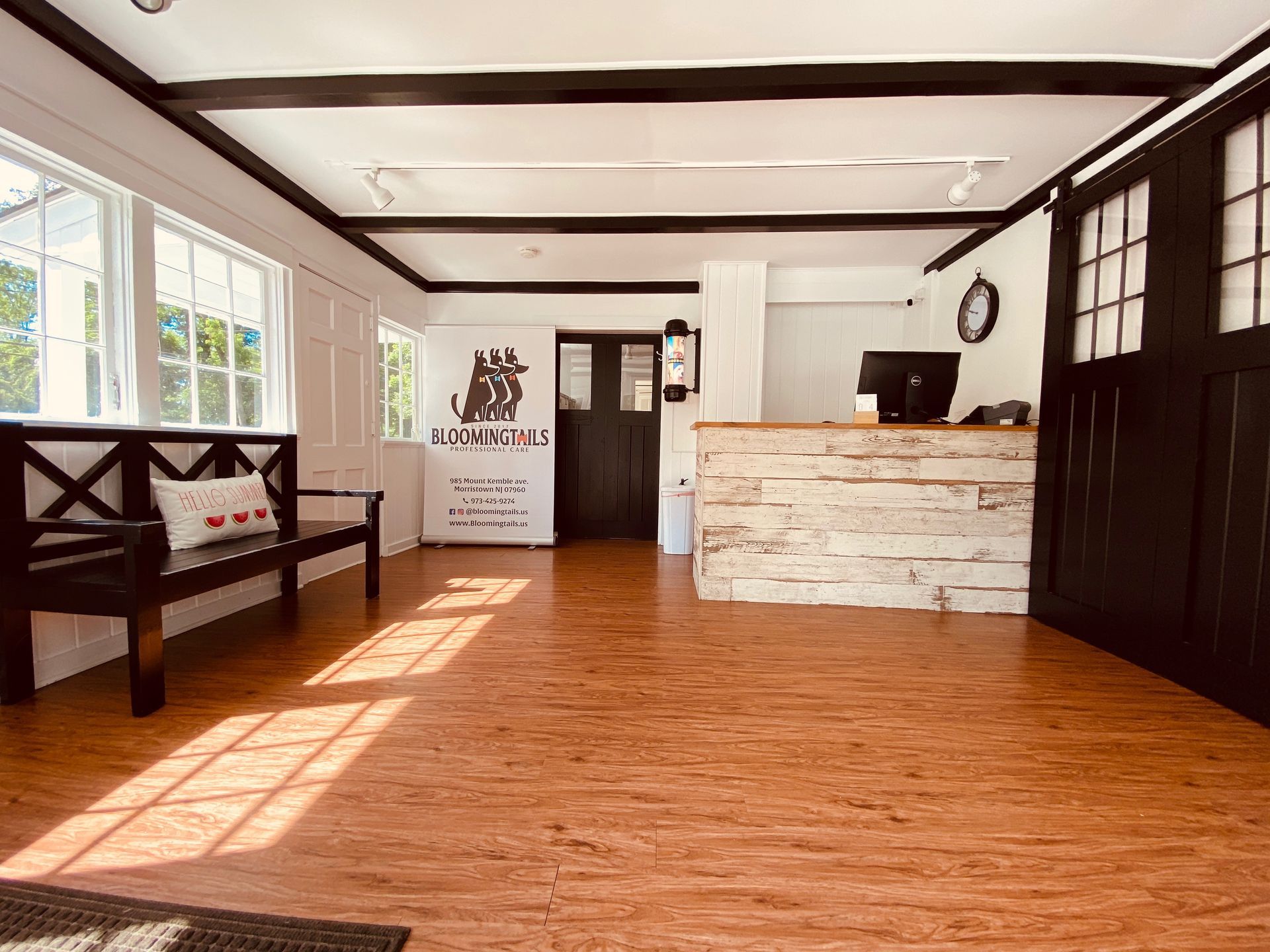 Bright reception area with wood floors, white walls, black trim, bench, and a wooden front desk near large windows.