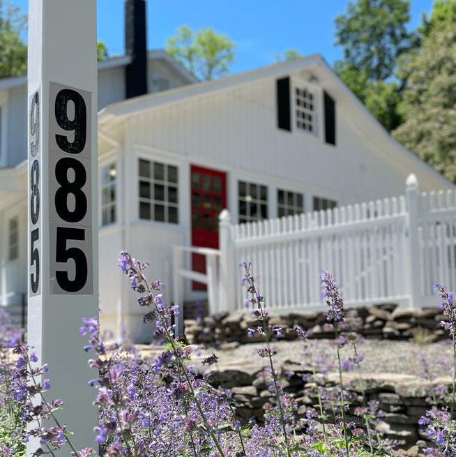White farmhouse with white fence and purple flowers, showing the number 985 on the front post.