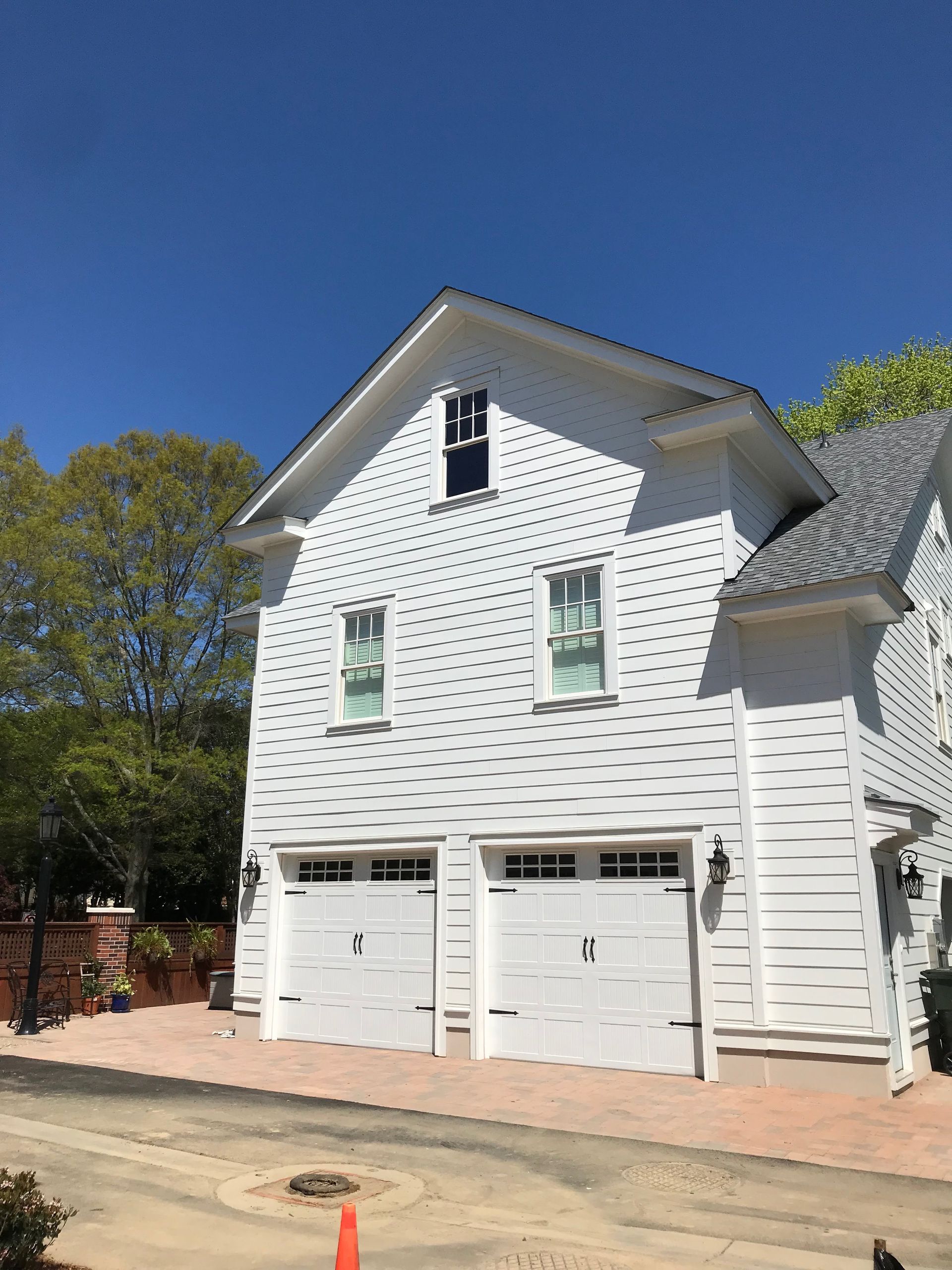 White two-story house with garage doors, multiple windows, and a gray roof on a sunny day.