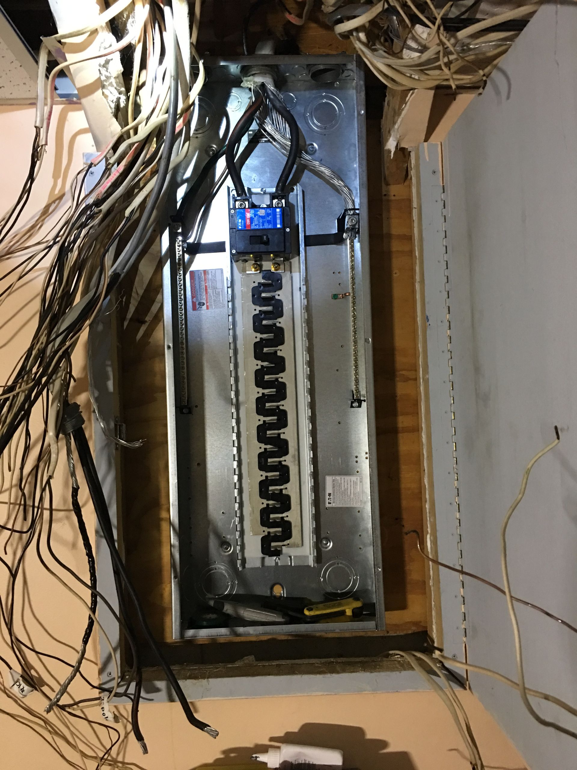 Exposed electrical panel with tangled wires and open breaker box in a damaged wall.