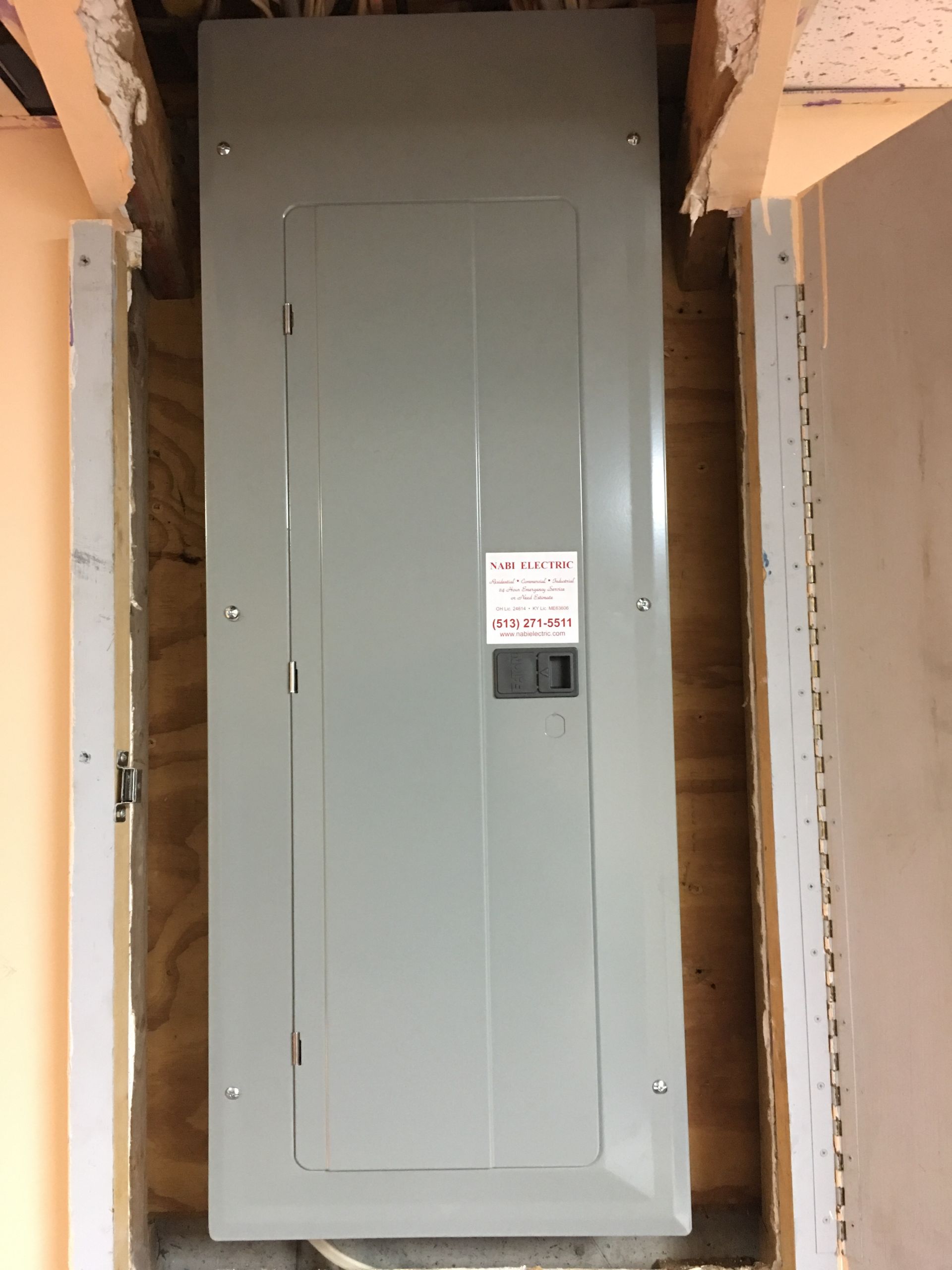 Gray electrical breaker panel mounted between exposed wall studs with a small warning label.