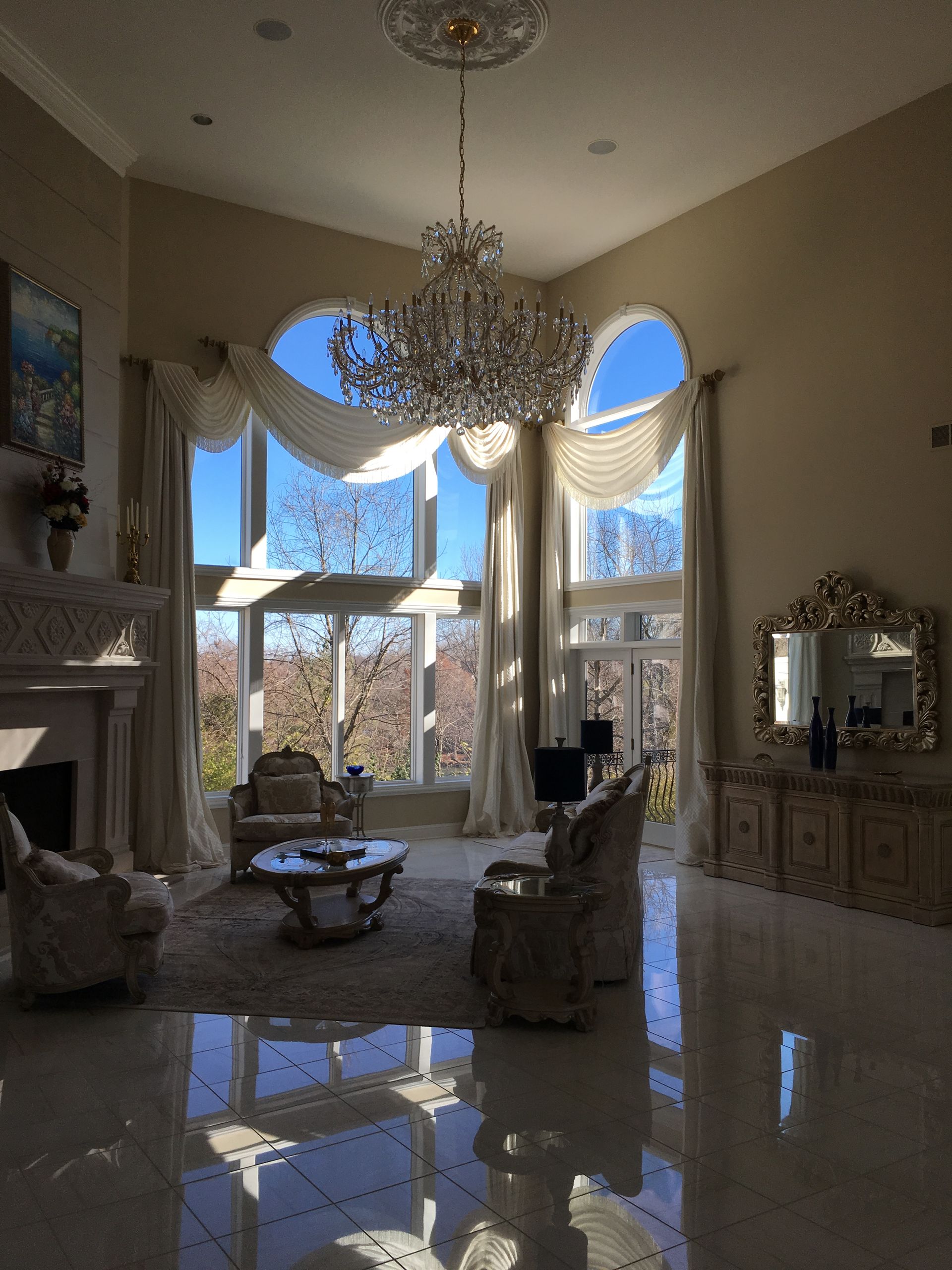 Grand living room with chandeliers, tall arched windows, and polished marble floors reflecting sunlight