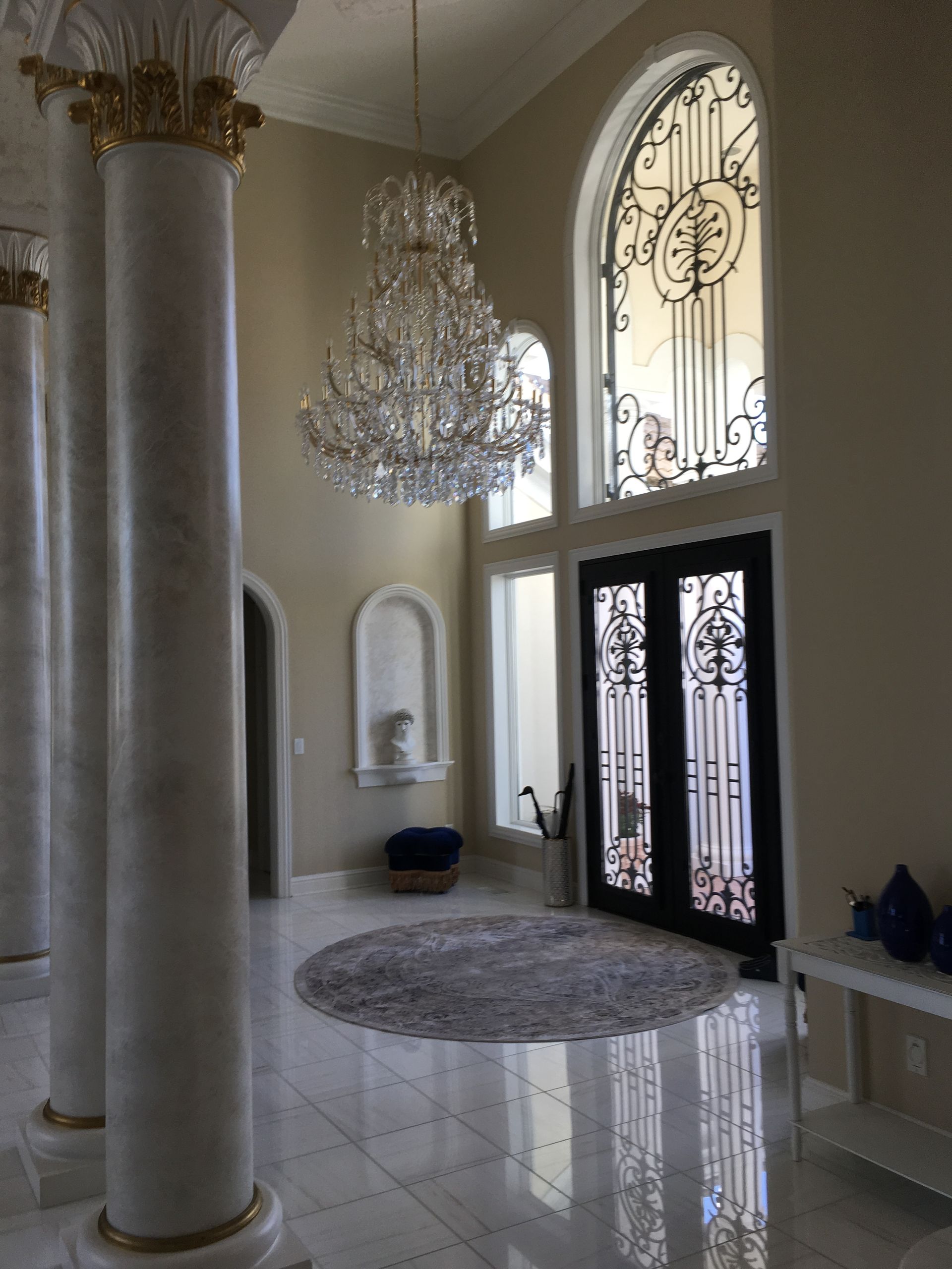 Elegant foyer with marble floor, crystal chandelier, tall columns, and ornate double doors beneath an arched window