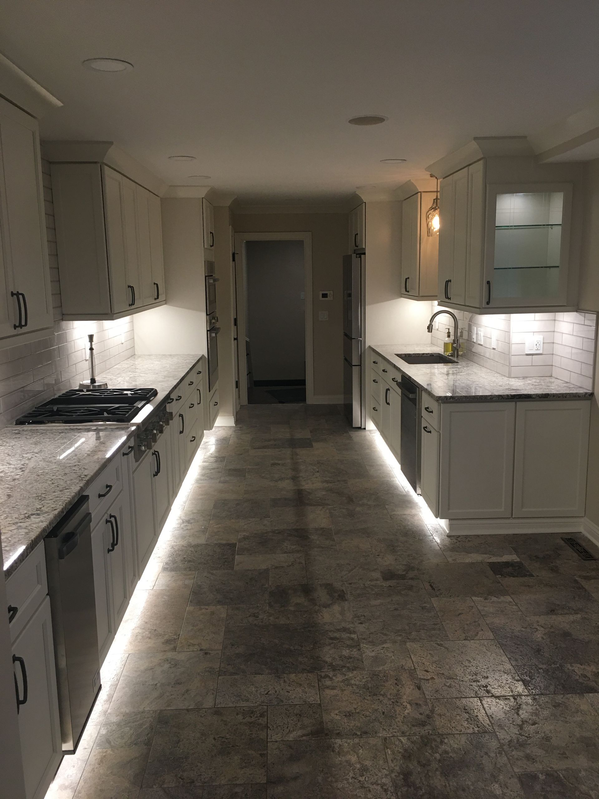 Long white kitchen with marble countertops, under-cabinet lighting, and a dark stone floor leading to a hallway