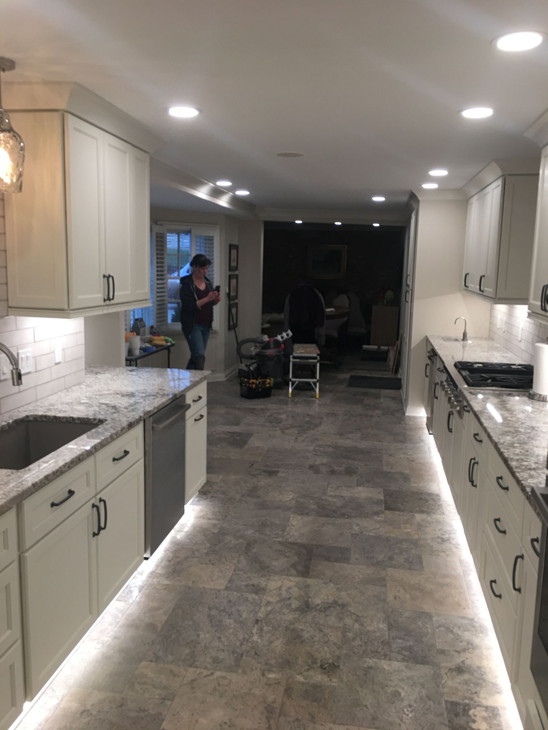 Bright kitchen with white cabinets, gray tile floor, and recessed lighting, leading to a dark room in back.