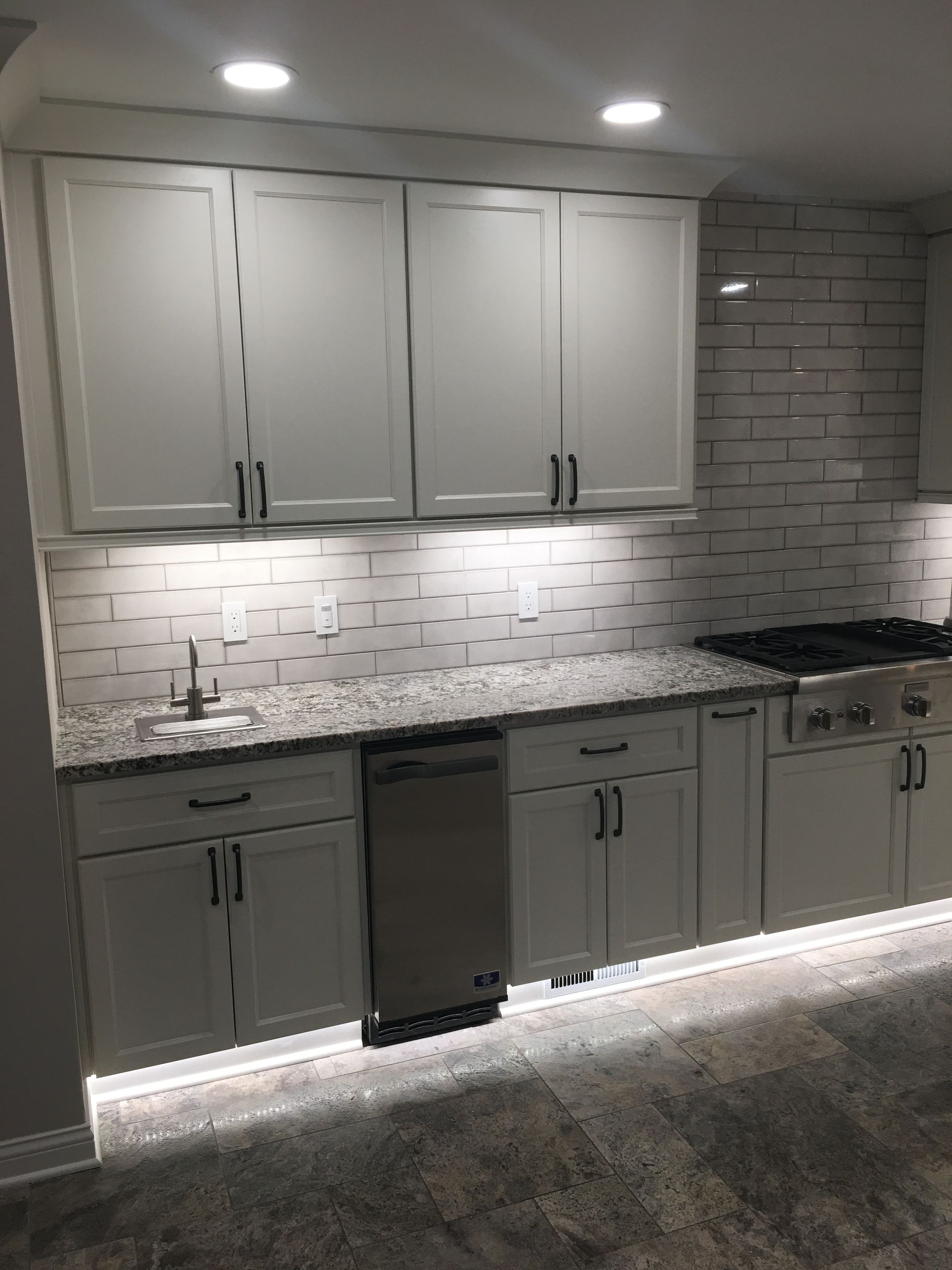 Modern kitchen with gray cabinets, white tile backsplash, granite counters, and under-cabinet lighting.
