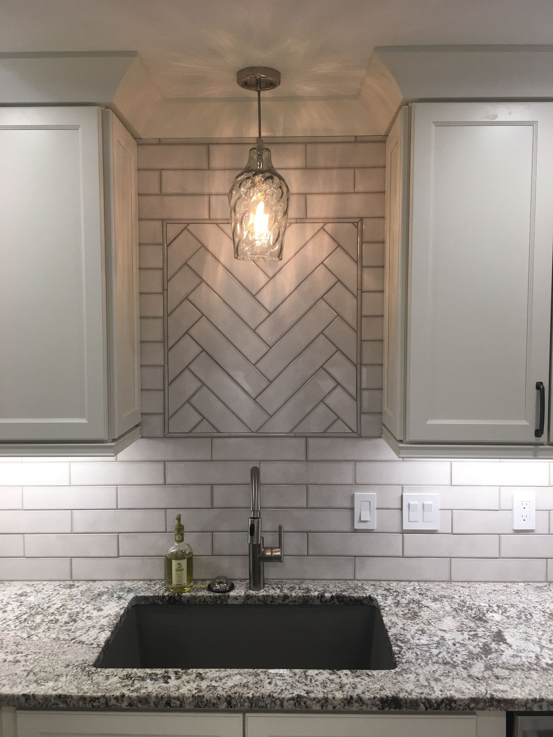 Kitchen sink beneath herringbone tile backsplash and pendant light, with white cabinets and granite countertop