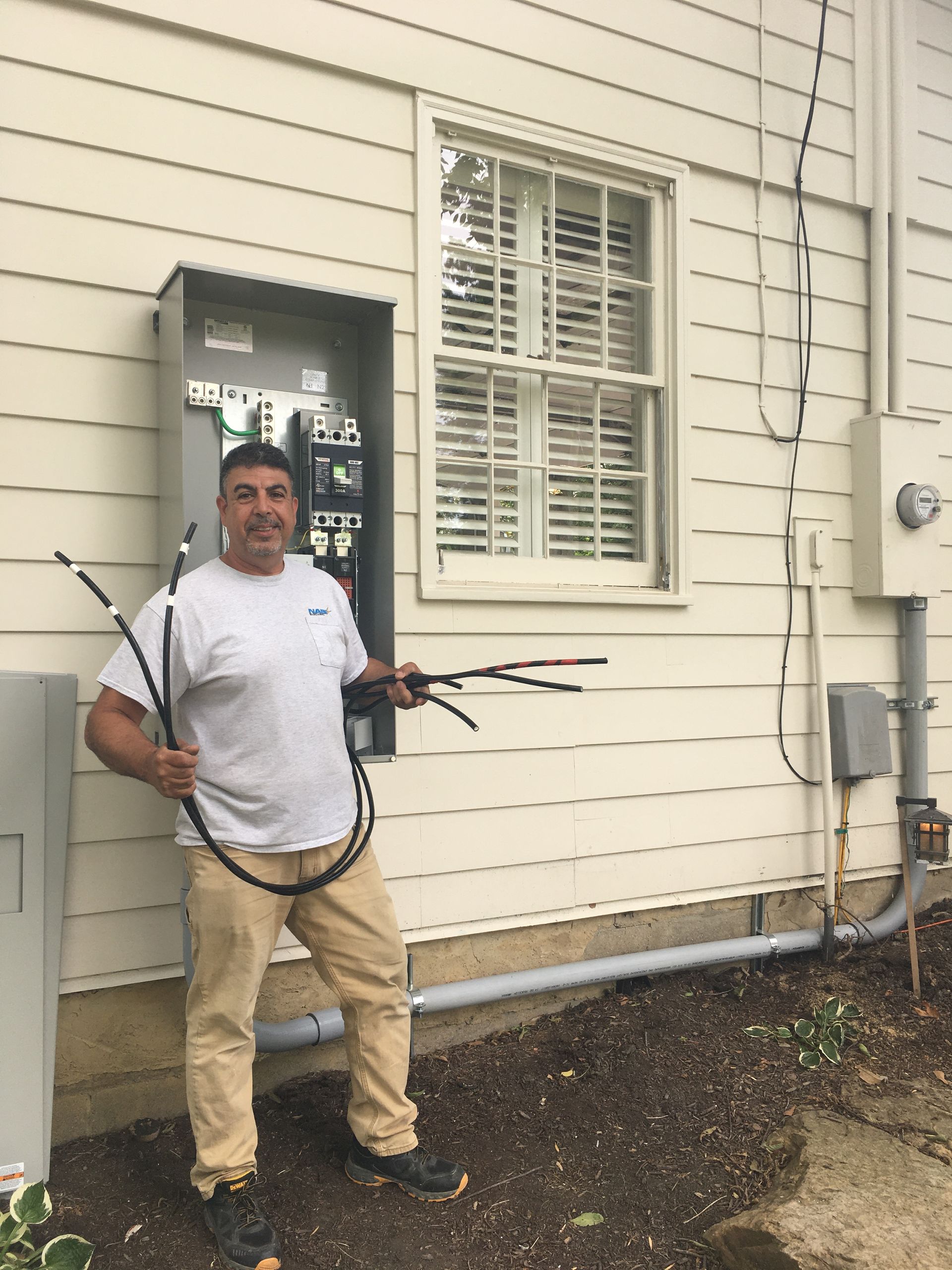 Man holding cables and tools beside a house exterior with window and utility boxes