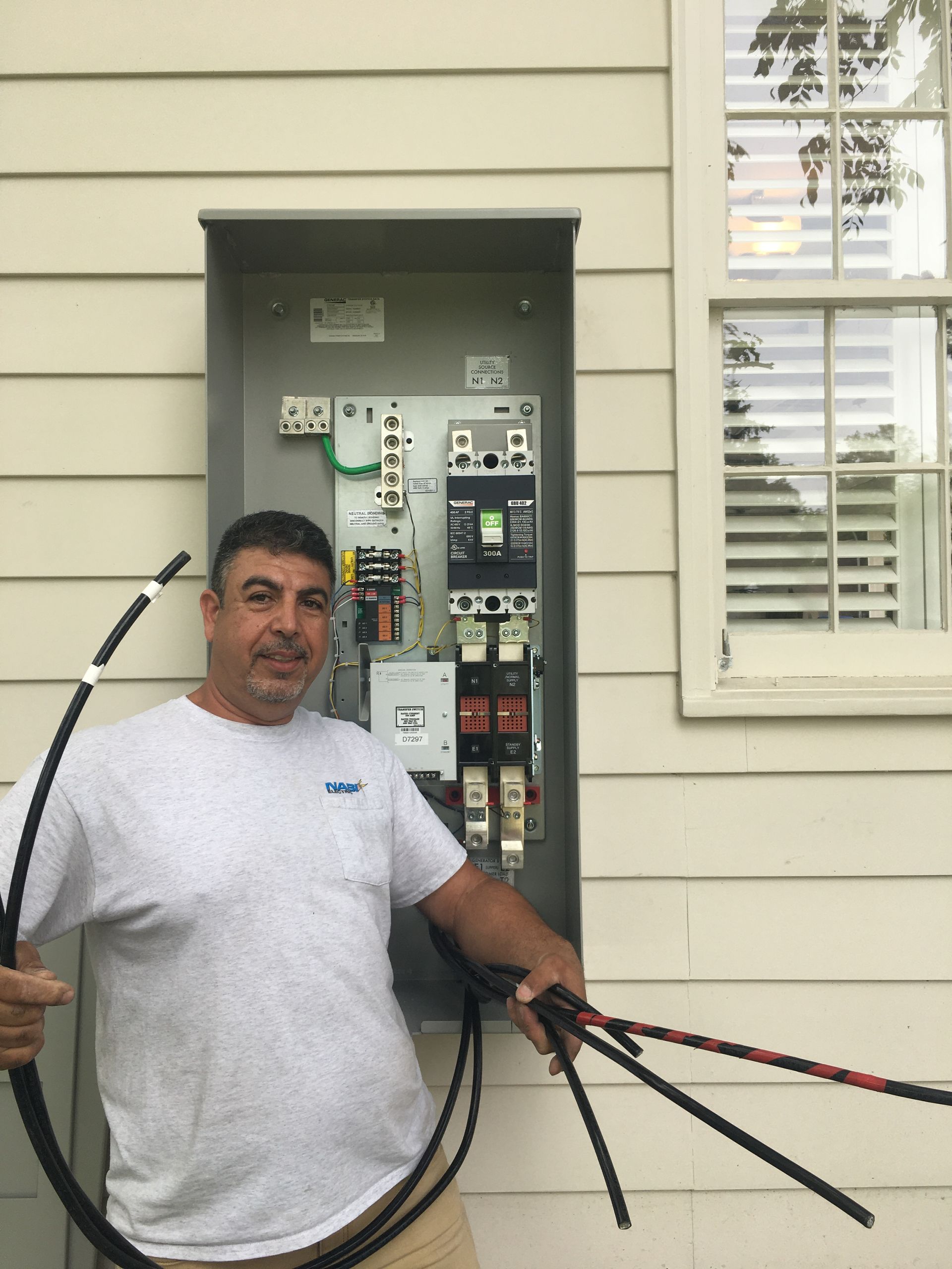 Man standing beside an open electrical panel holding black cables outside a house
