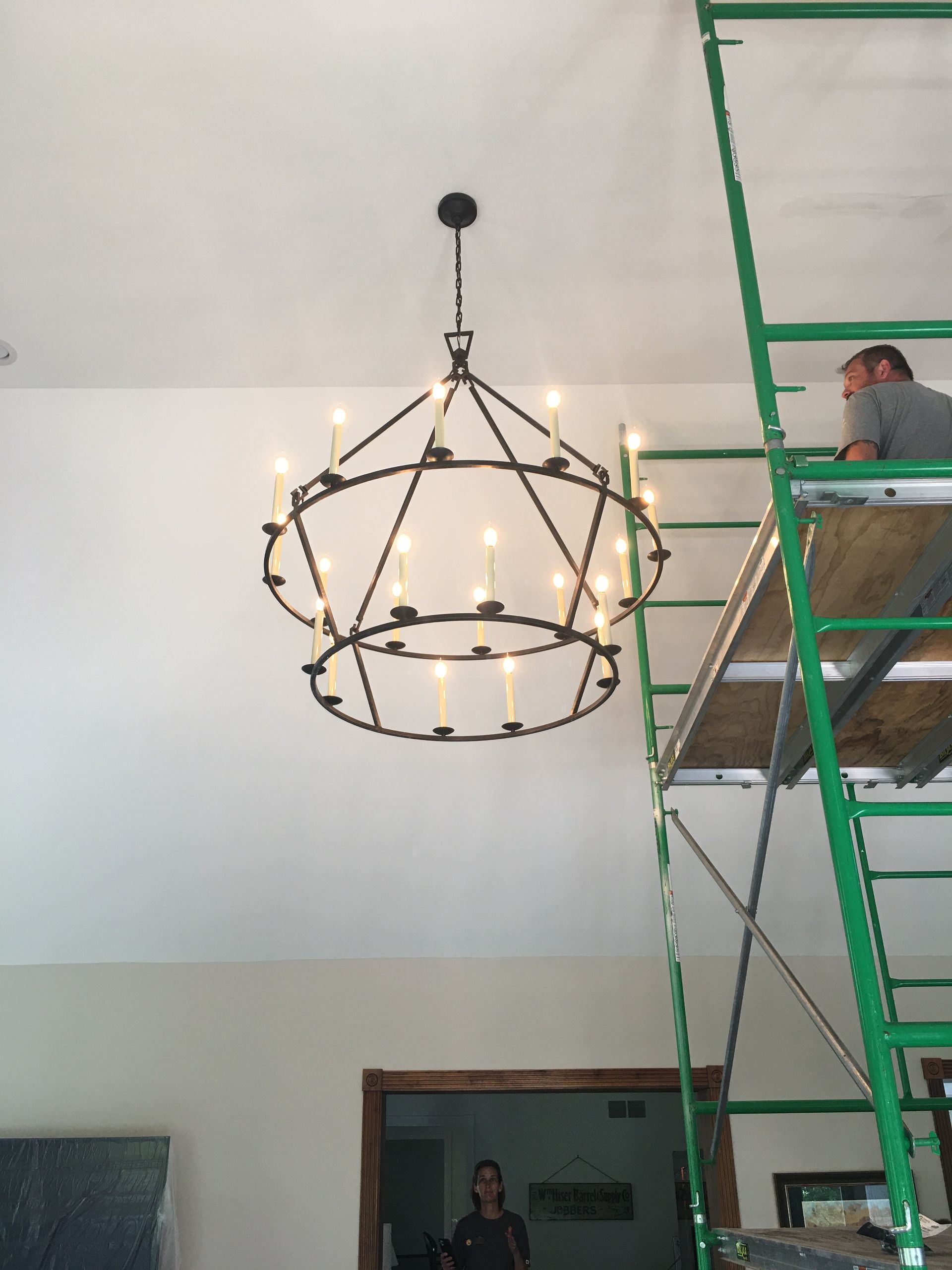 A chandelier being installed in a room, with a worker on scaffolding to the right.