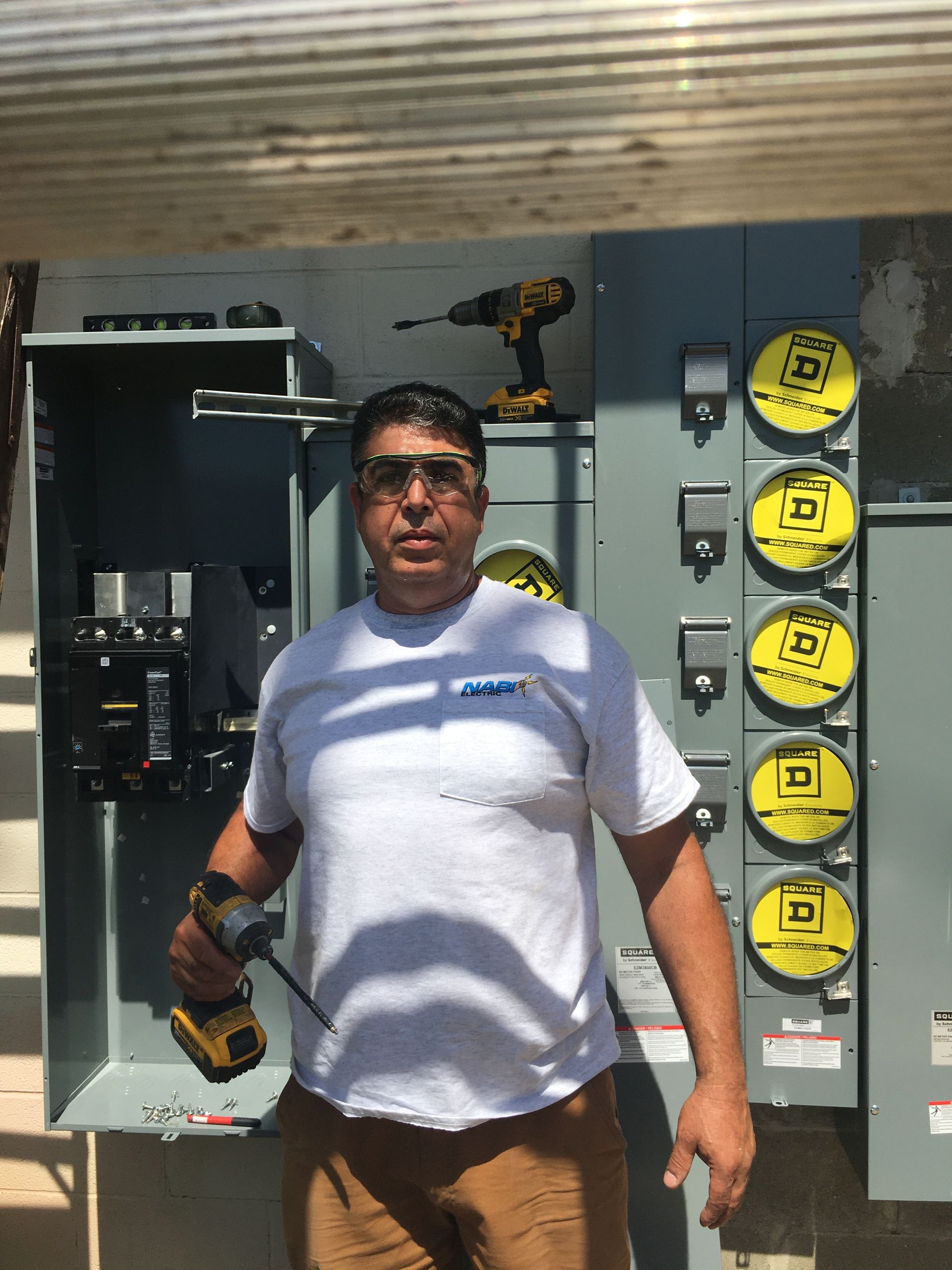 Worker holding a power drill stands in front of an open electrical panel outdoors.