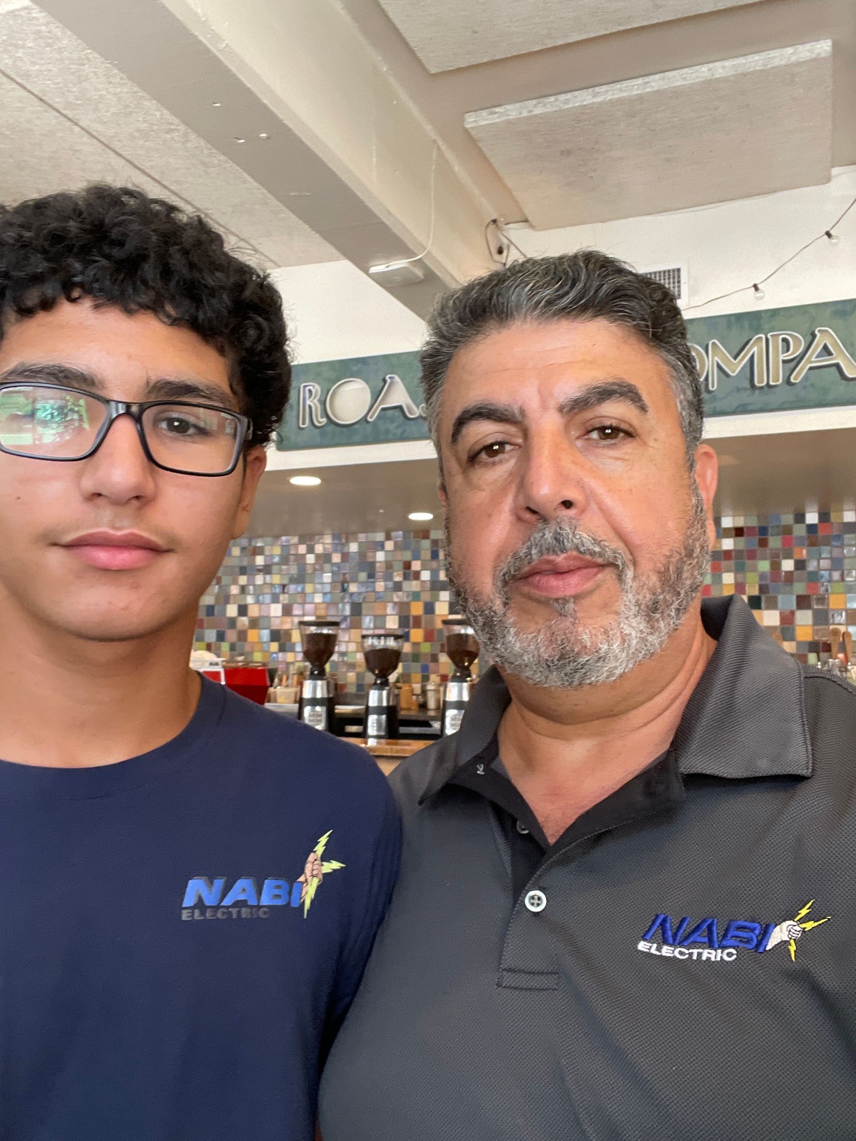 Two people standing indoors, wearing matching navy shirts, in front of a shop counter and product display.