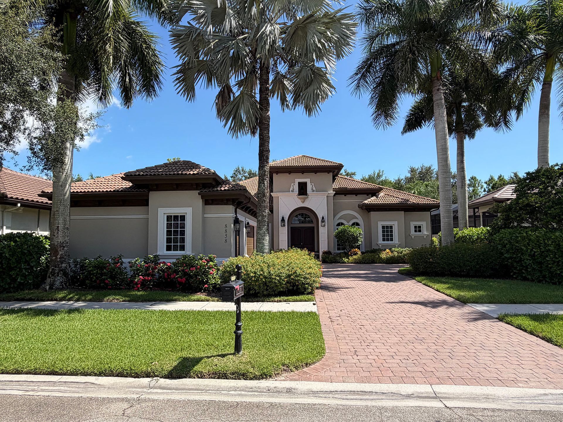 Mediterranean-style house with palm trees, landscaped front yard, and brick driveway in bright sunlight