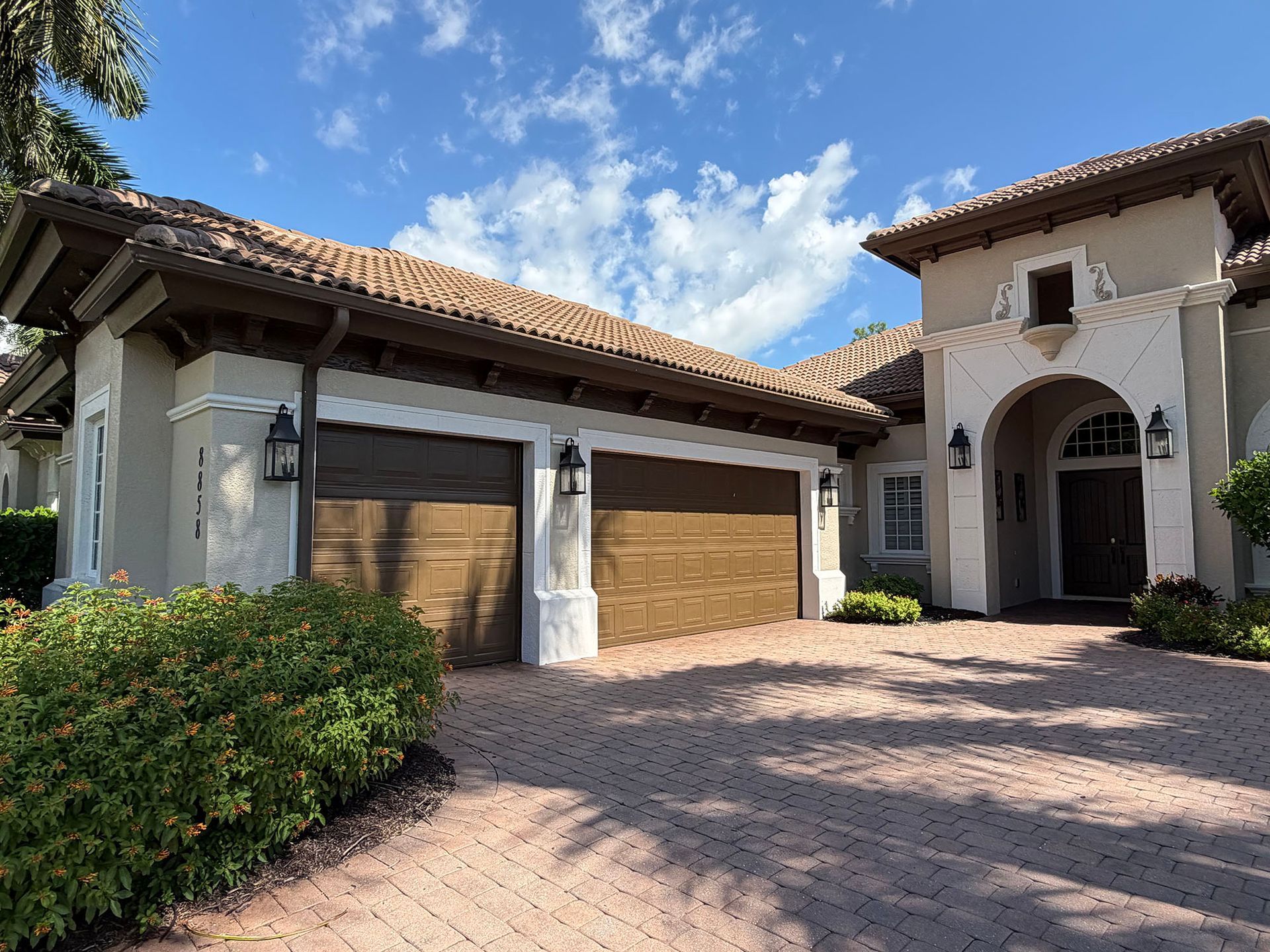 Mediterranean-style house exterior with two wooden garage doors, arched entryway, and a brick driveway under a blue sky