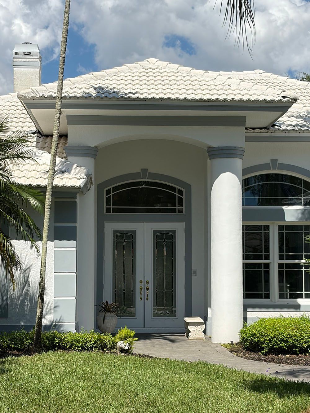White house front entrance with arched windows, columns, and a tiled roof behind a green lawn.