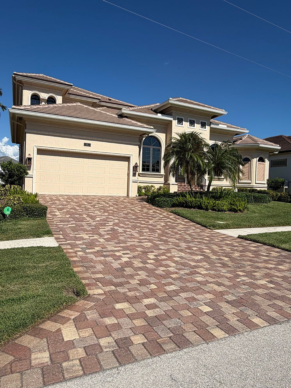 Suburban stucco house with a brick driveway, garage, and manicured front lawn under a clear blue sky