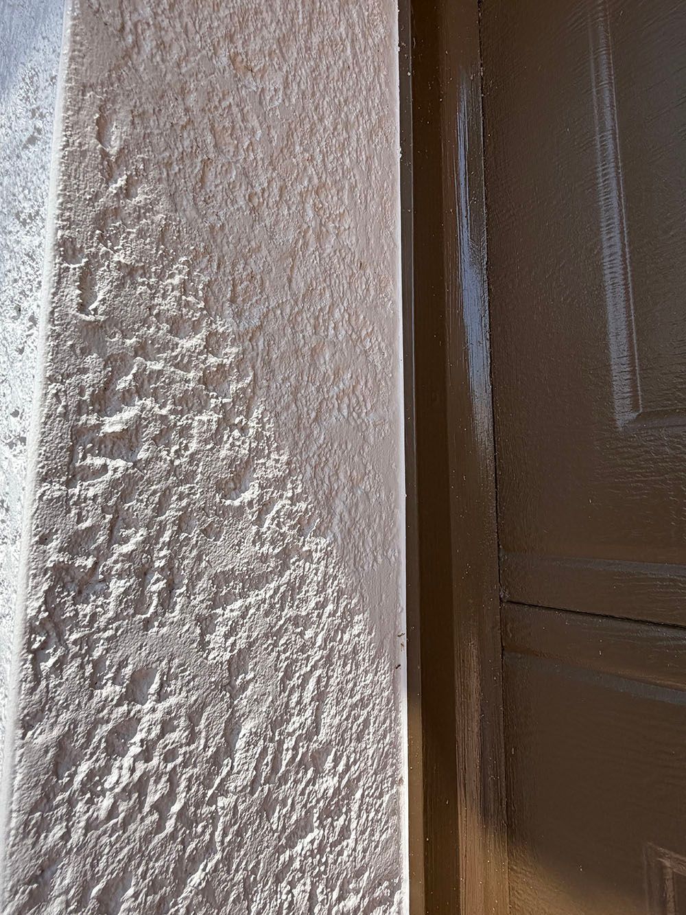 Close-up of a beige stucco wall beside a brown door frame in daylight