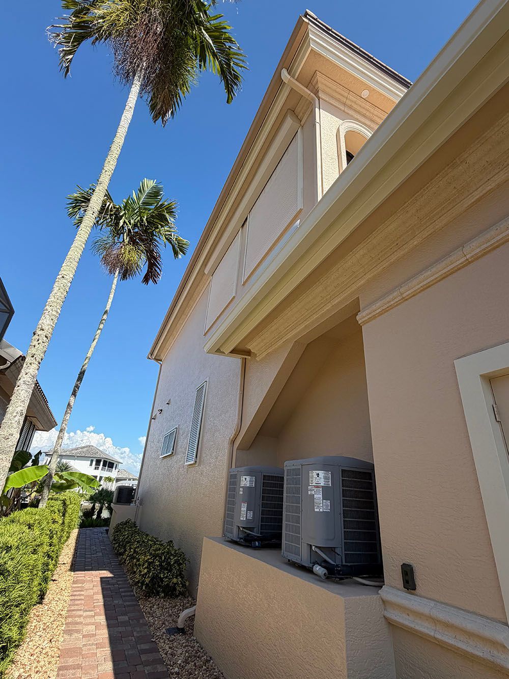 Tan stucco house exterior with tall palm trees and two air-conditioning units by a hedge walkway