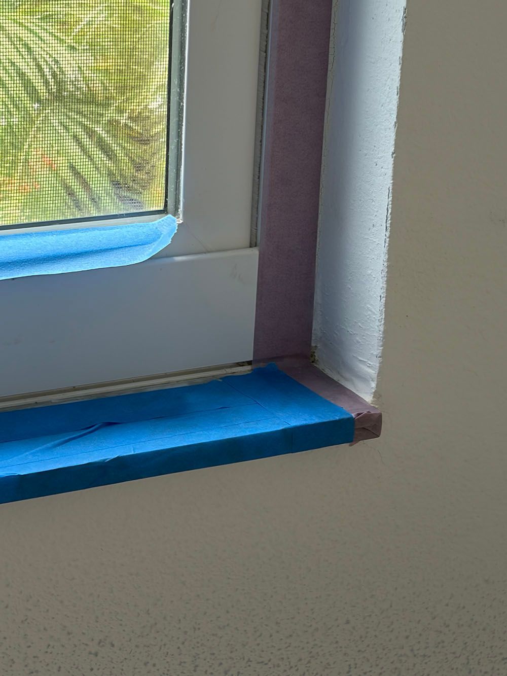 Close-up of a white windowsill with bright blue tape and a mesh screen beside a wall corner.