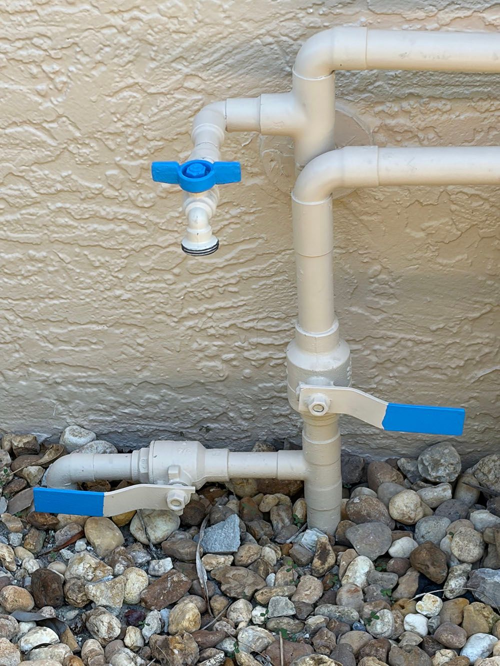 Outdoor water pipes with blue-handled valves against a beige wall above gravel.