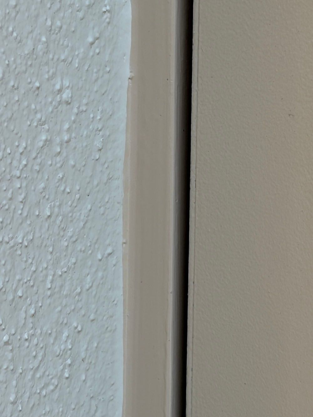 Close-up of a narrow gap between two painted wall or door surfaces, one textured white and one smooth beige.